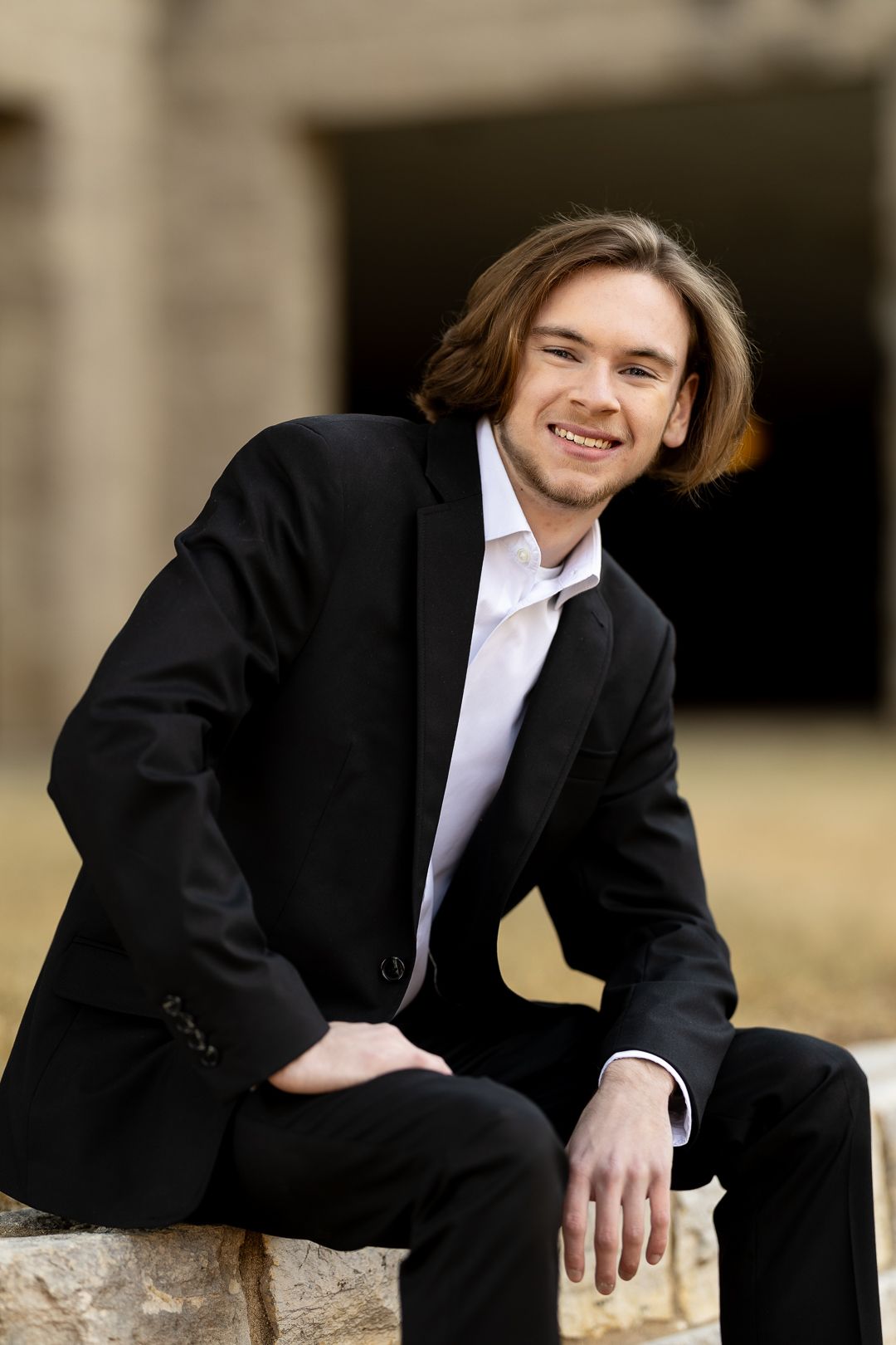A person in a black suit jacket and white shirt sits casually on a stone ledge outdoors.
