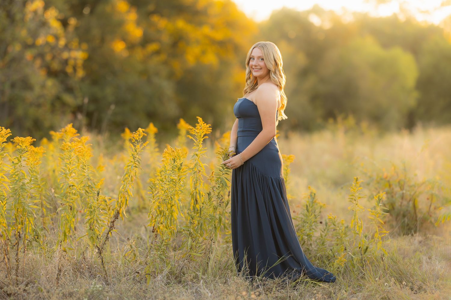A person in a blue dress walks through a golden field at sunset during a dreamy outdoor photo session.