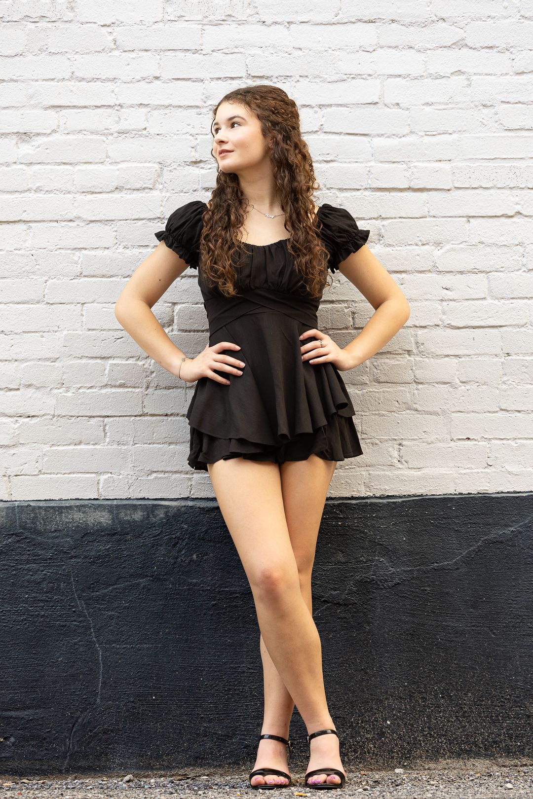 Woman in black ruffled top poses against white brick wall in artistic portrait series.