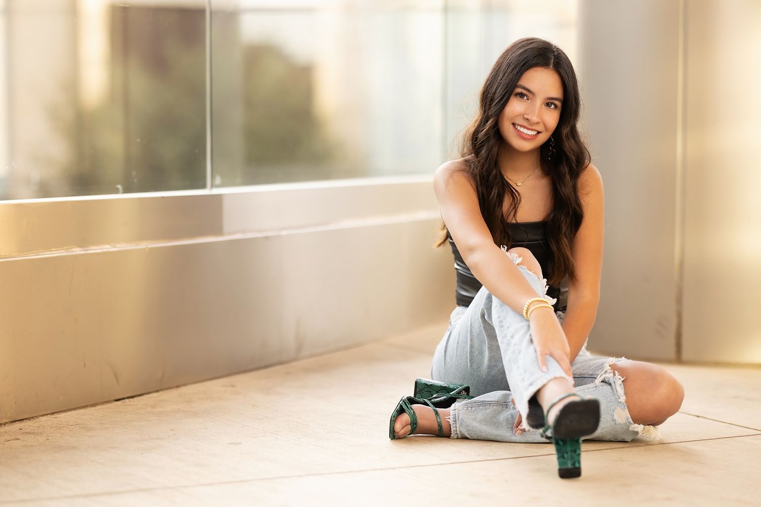 A woman in casual attire smiles while sitting near a modern glass wall with natural lighting.