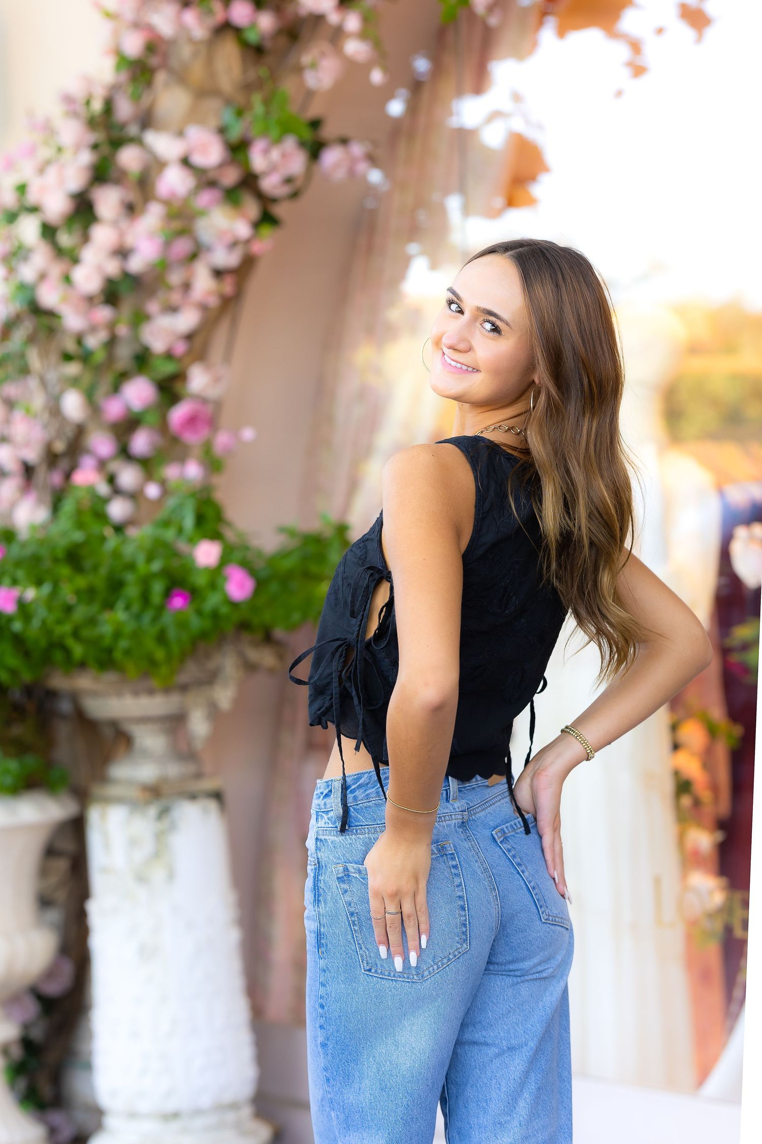 A person poses in denim overalls and a black tank top against a backdrop of pink flowering vines.