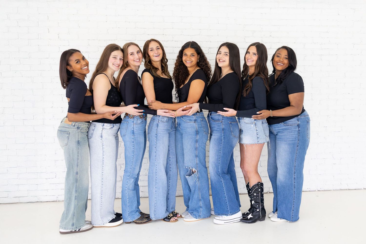 A group of friends wearing denim jeans and casual outfits pose together against a white wall.