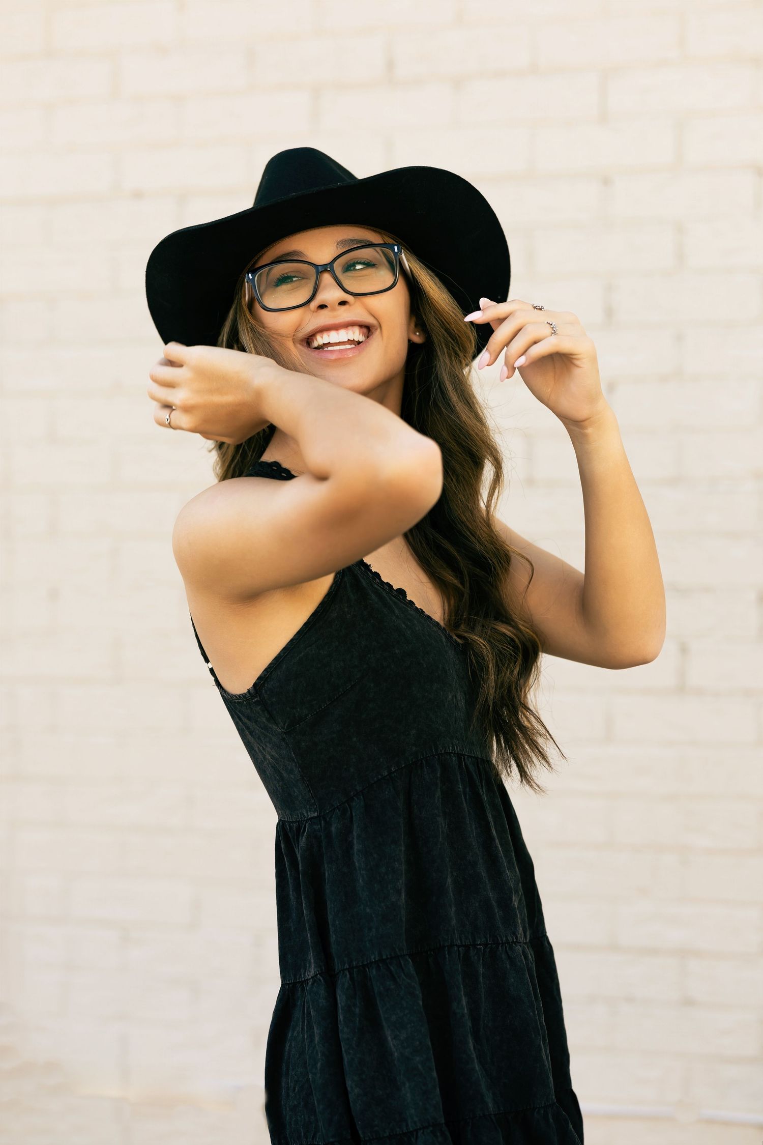 A joyful person wearing a black sundress and wide-brimmed hat poses against a white brick wall.