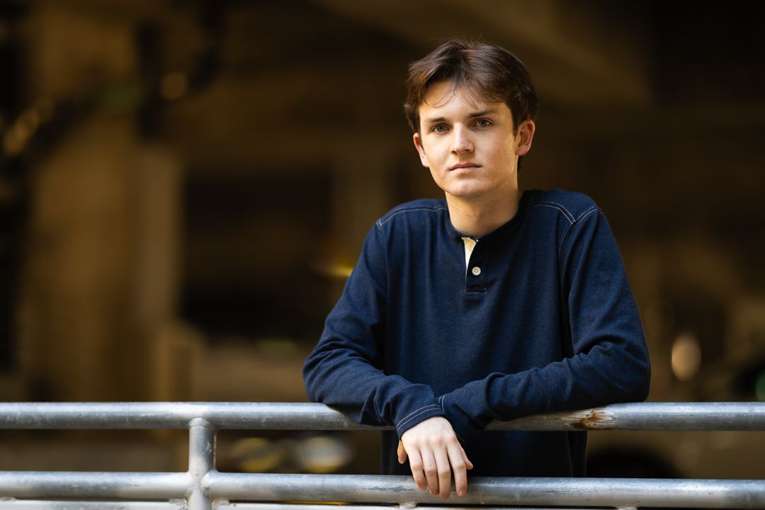 A person in a navy blue sweater leans against a railing in a dimly lit indoor setting.