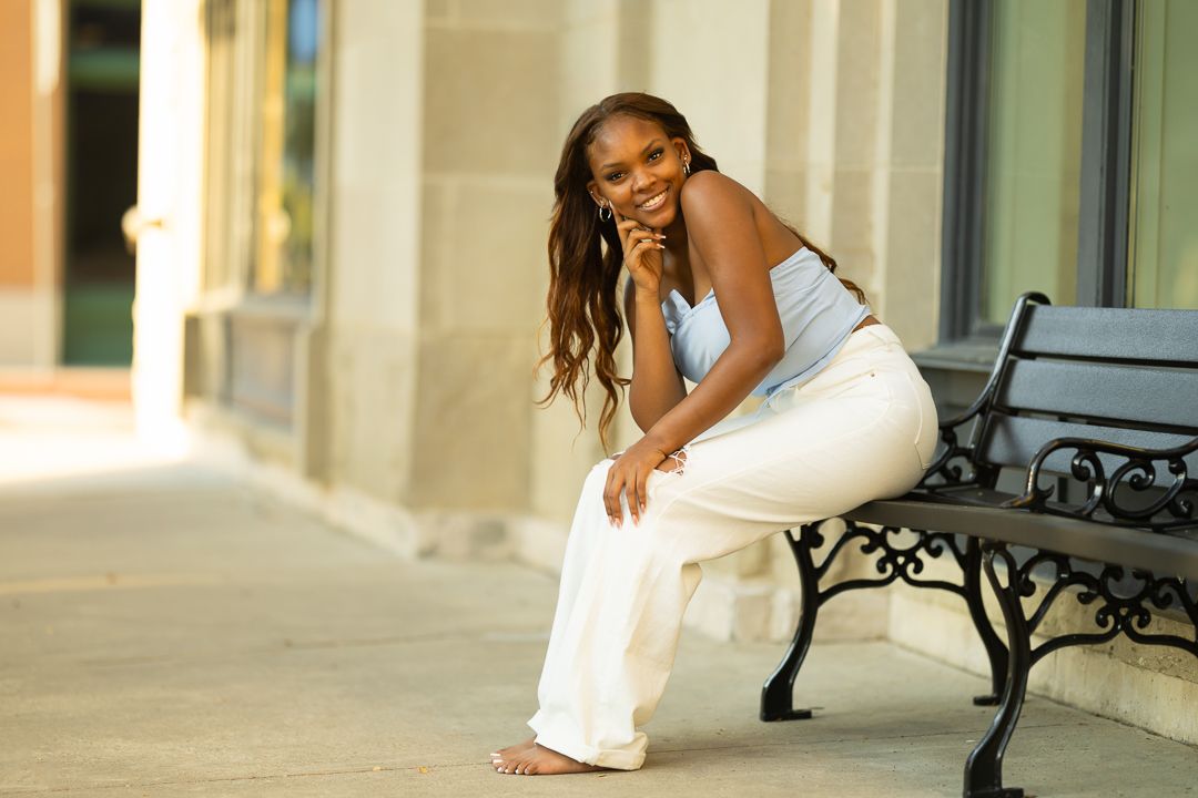 A person in a white dress poses playfully on an ornate wrought iron bench against a light-colored building exterior.