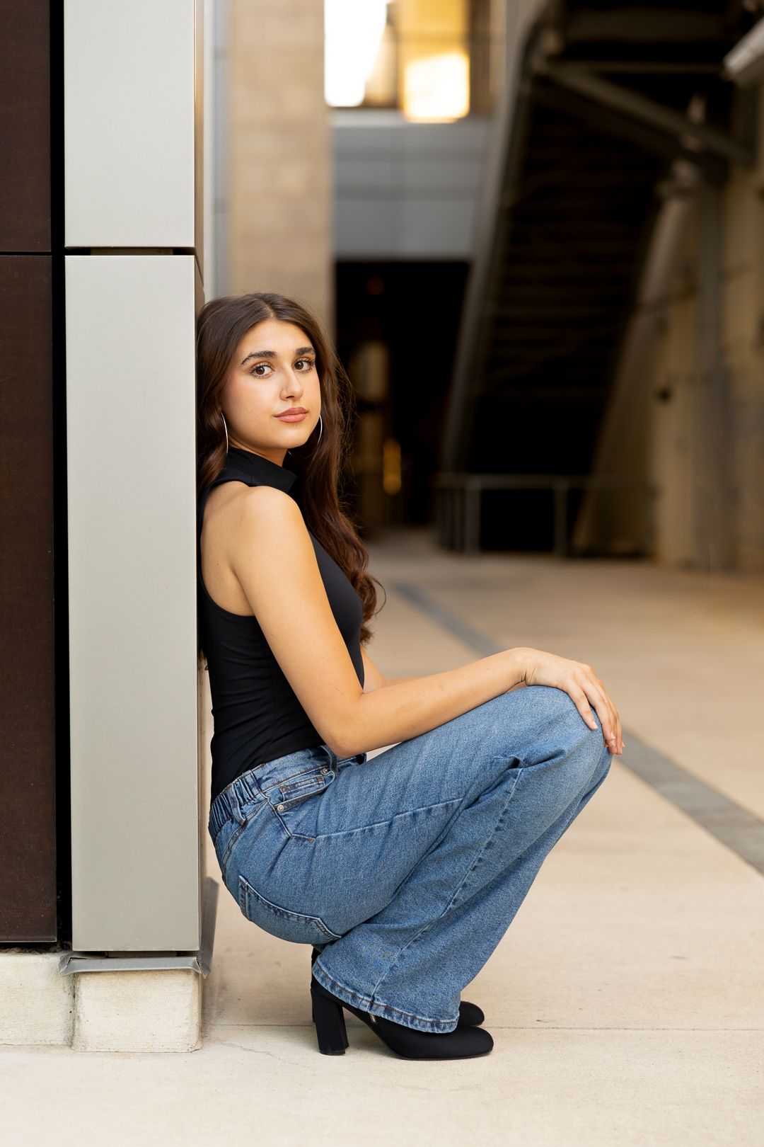 A person in a black tank top and wide-leg jeans crouches against a wall in an urban setting.