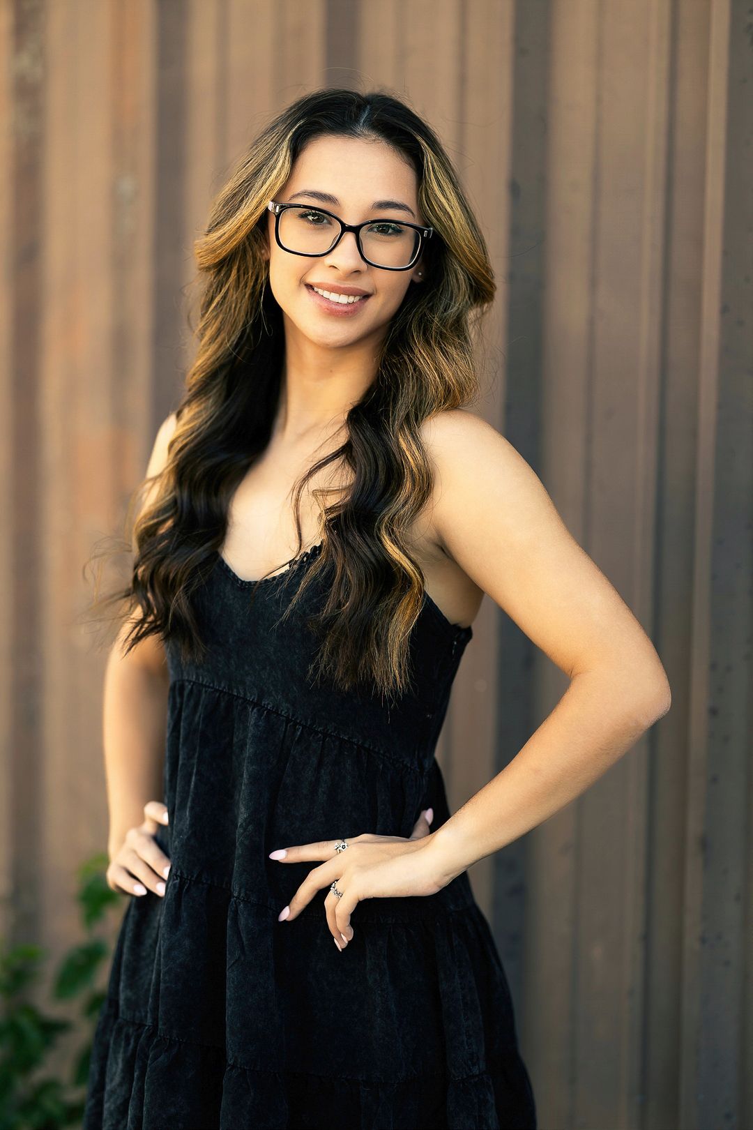 Stylish poses in a black sleeveless dress against a metallic wall background.