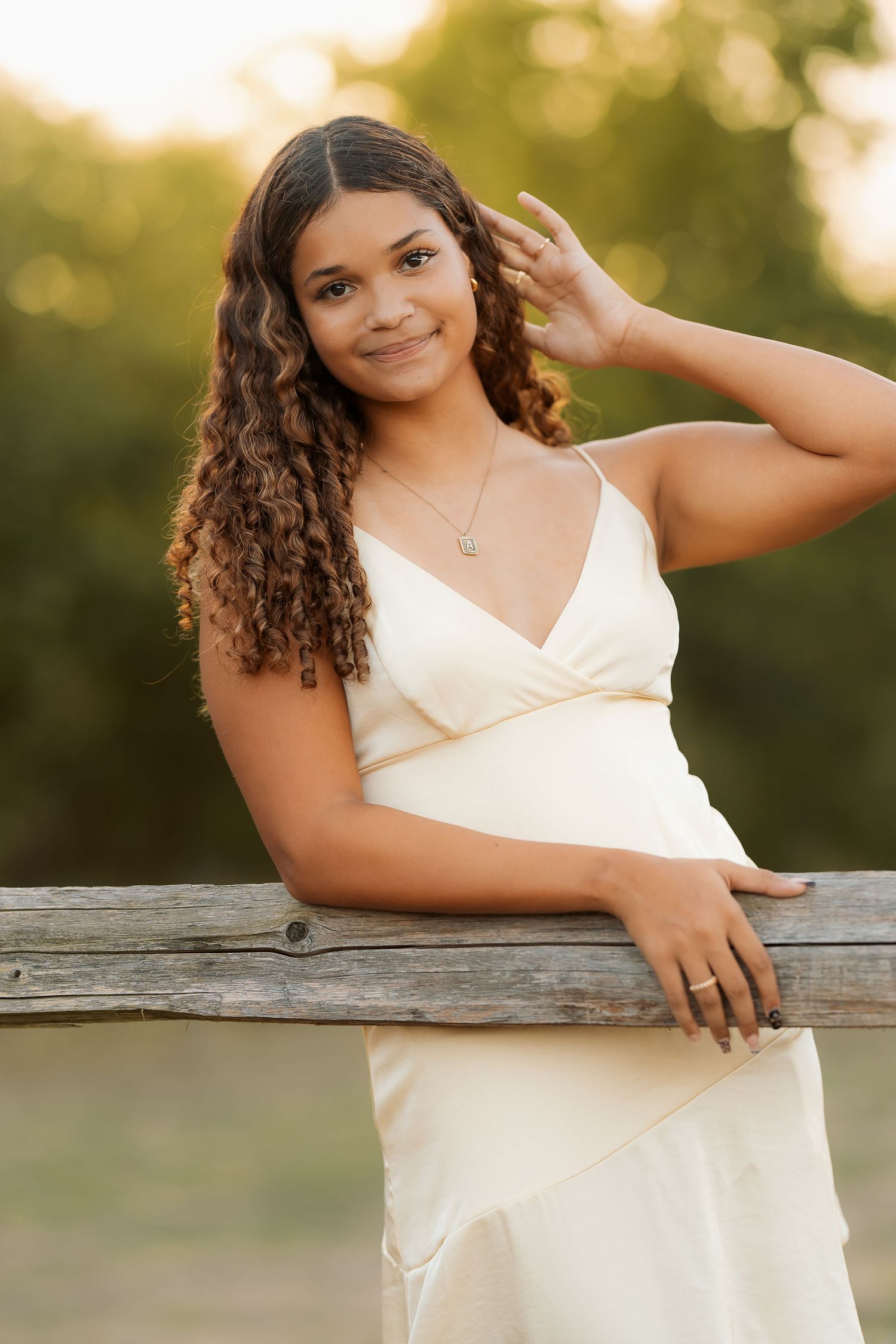 A woman in a cream dress poses by a wooden fence during golden hour.