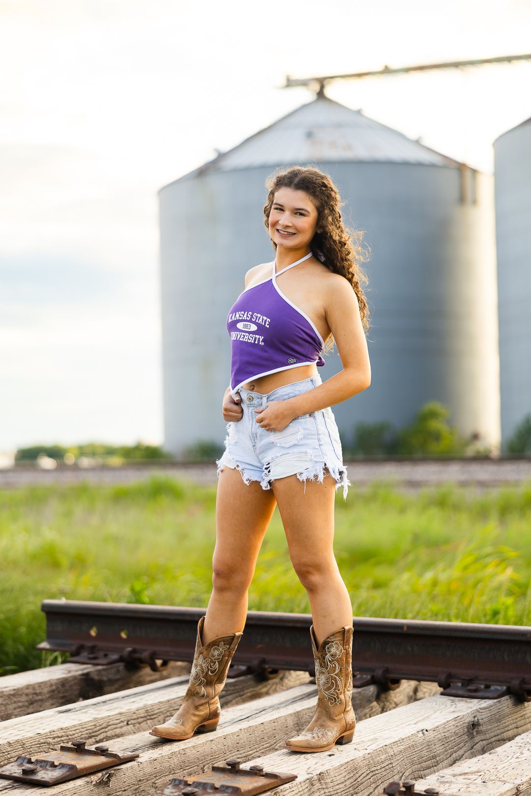 Woman in cowboy boots and denim shorts poses near railroad tracks with grain silos in the background.