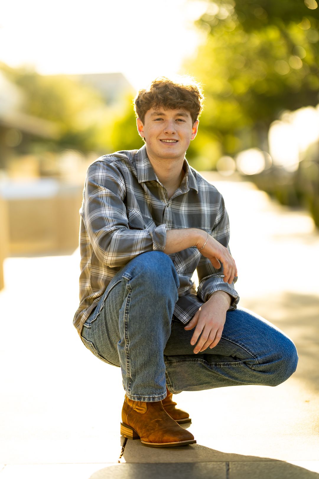 A person in a plaid shirt and jeans sits casually outdoors during golden hour with warm sunlit bokeh in the background.