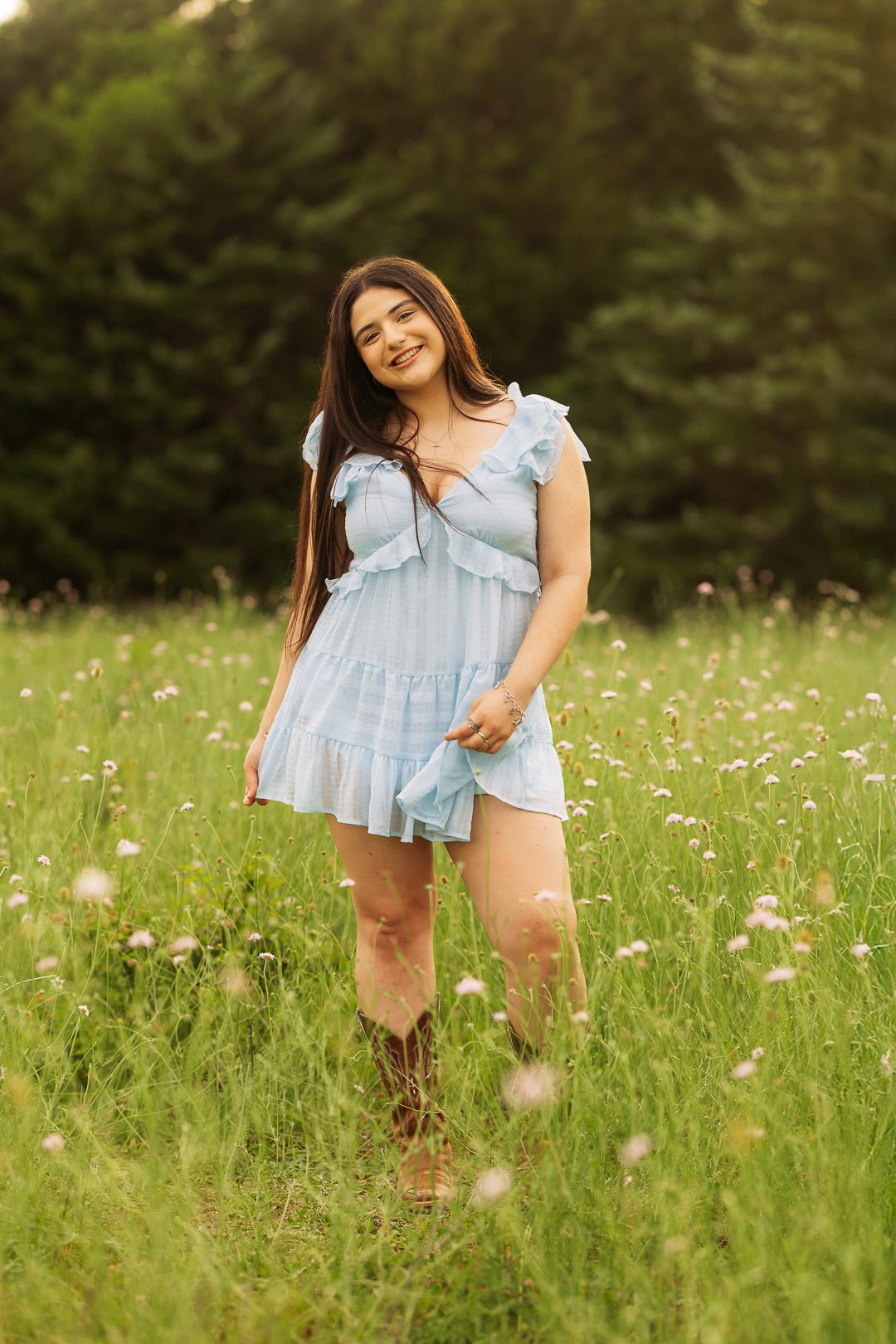 Young woman in light blue ruffled dress and cowboy boots smiling in a wildflower meadow with green trees behind her.
