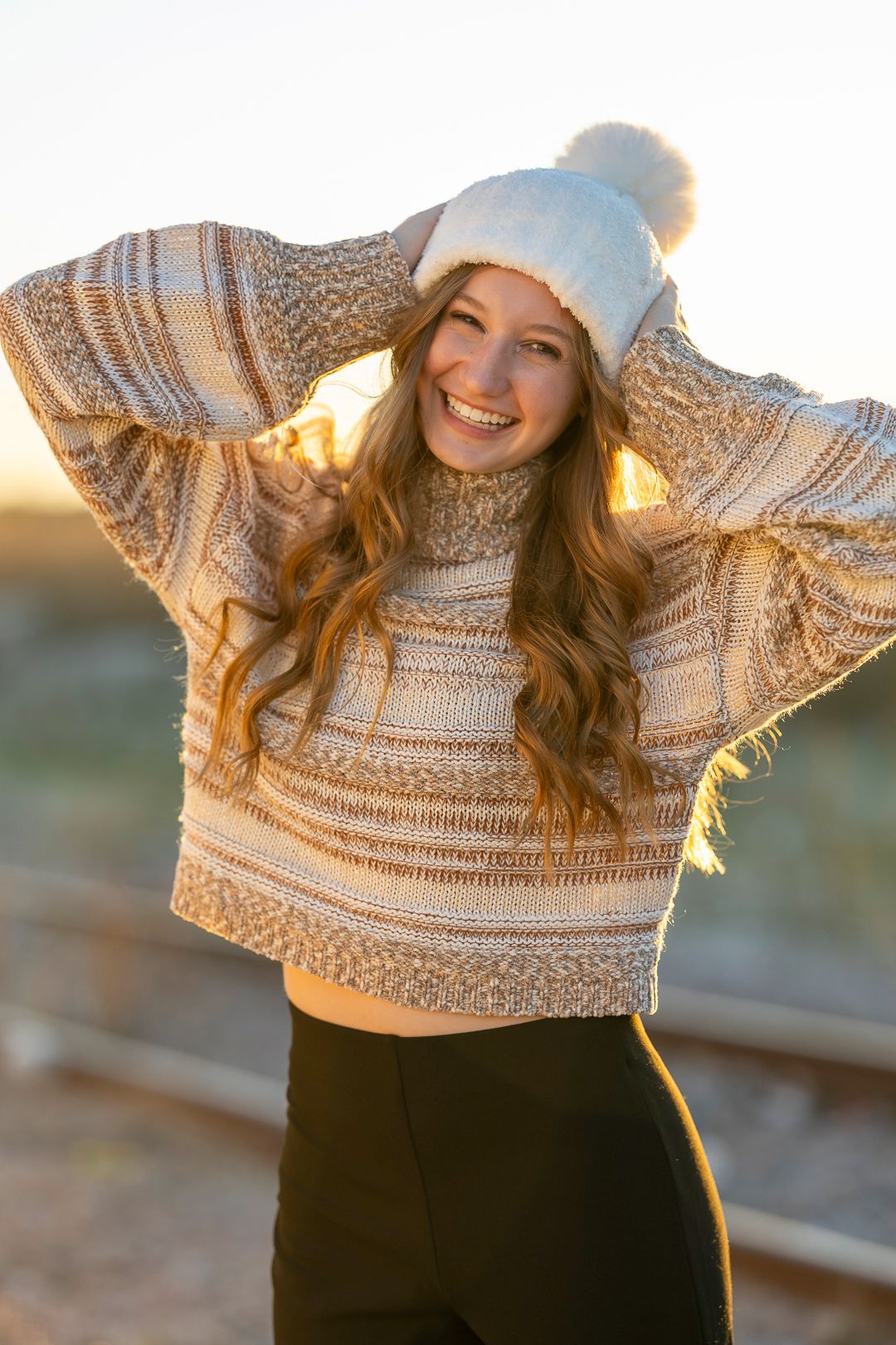 Person wearing cozy striped sweater and winter hat poses outdoors at golden hour.