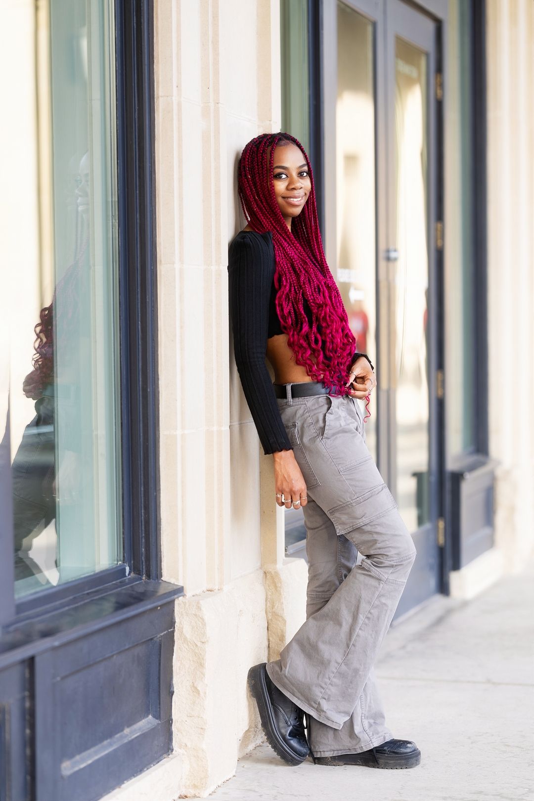 A person with vibrant red hair poses against a wall in a long gray dress and black boots.