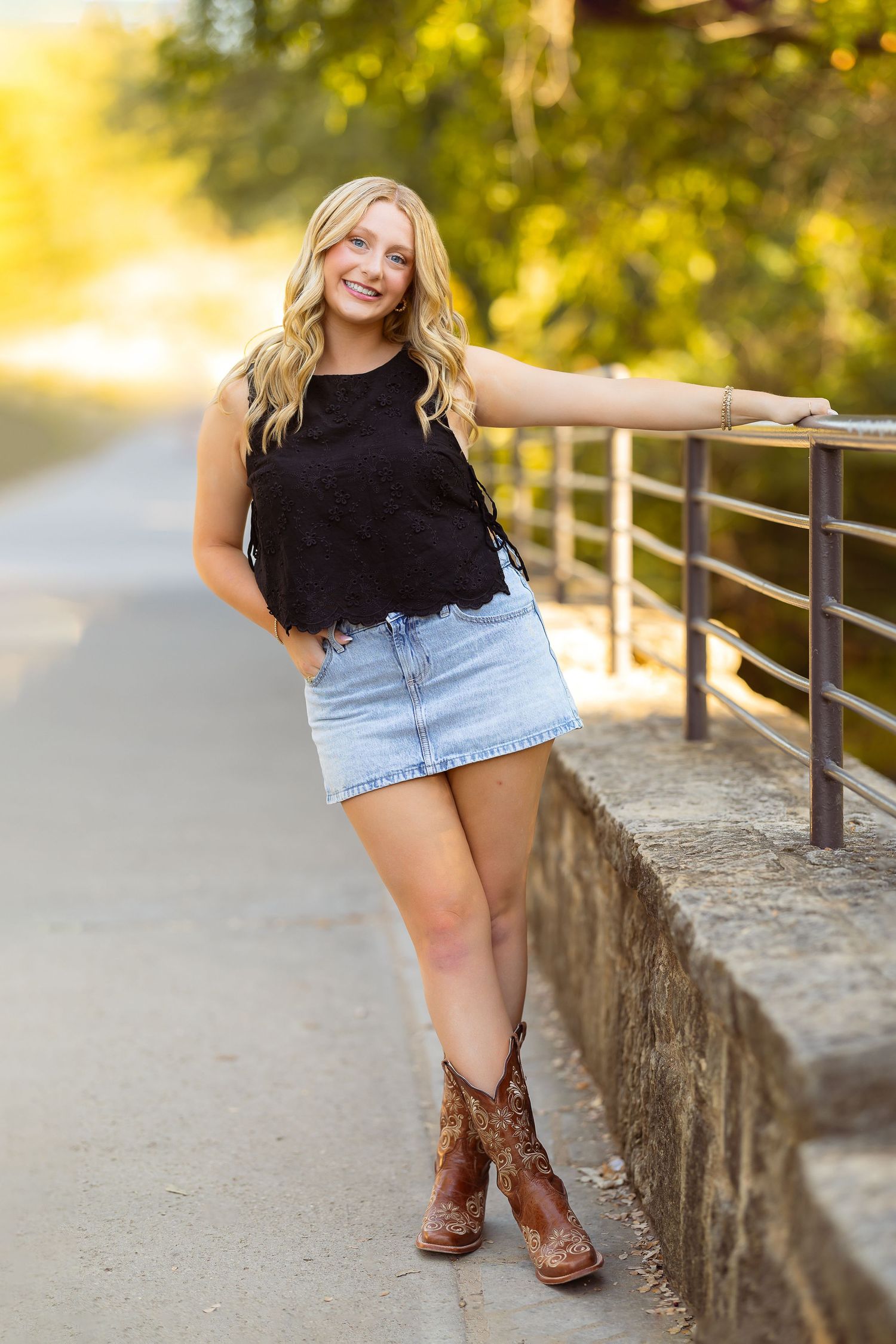 A person in a black top and denim shorts poses along a stone wall at sunset wearing brown cowboy boots.