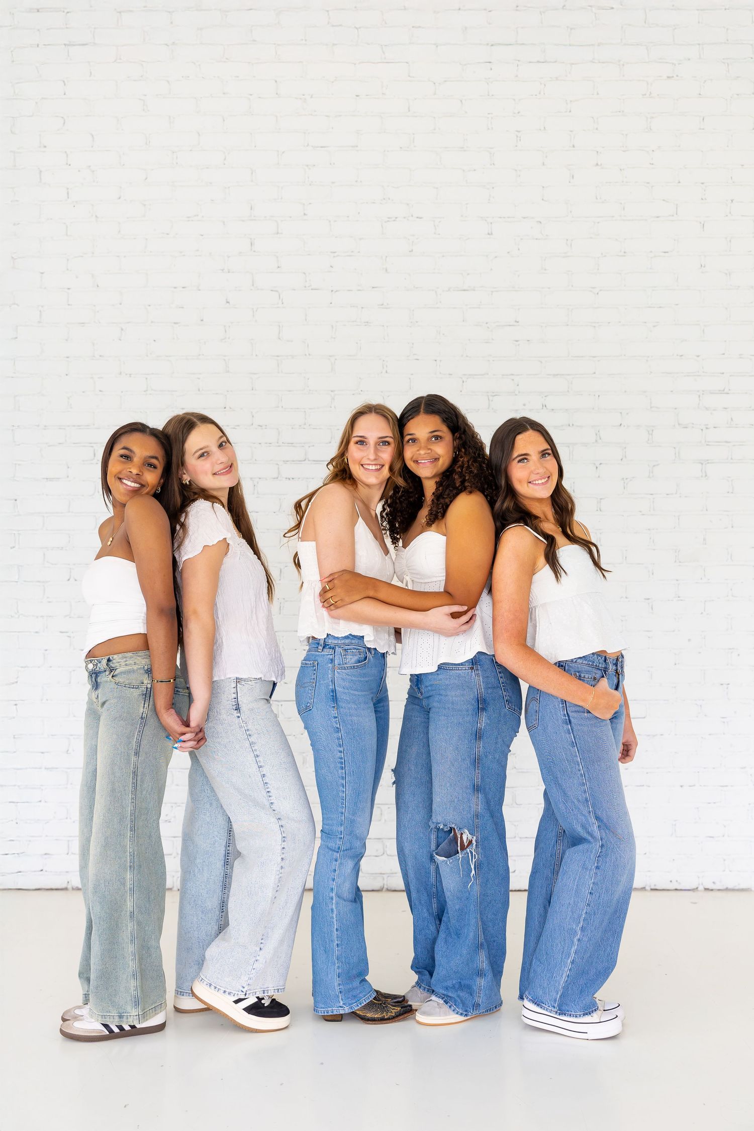 Group of friends in matching denim jeans and white tops pose together against a white wall.