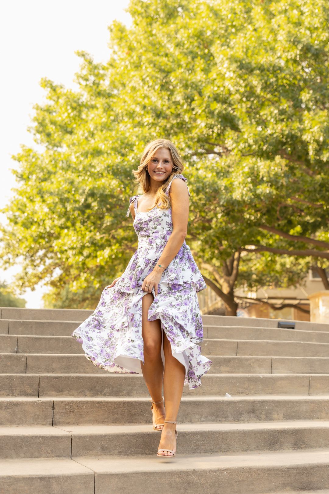 A person in a floral dress poses playfully on concrete steps in bright sunlight.