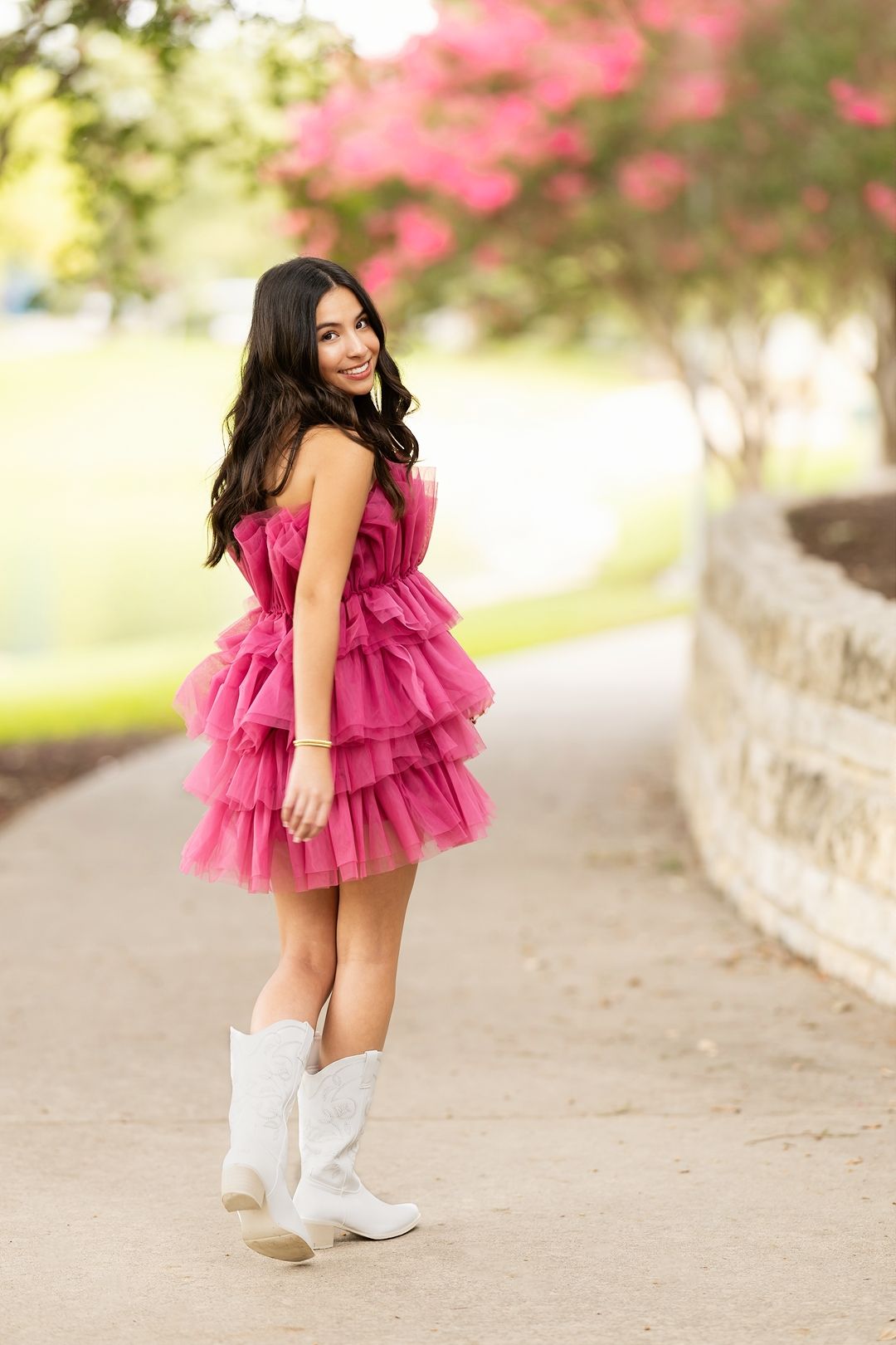 A child in a pink ruffled dress and white cowboy boots poses outdoors against a blurred floral background.