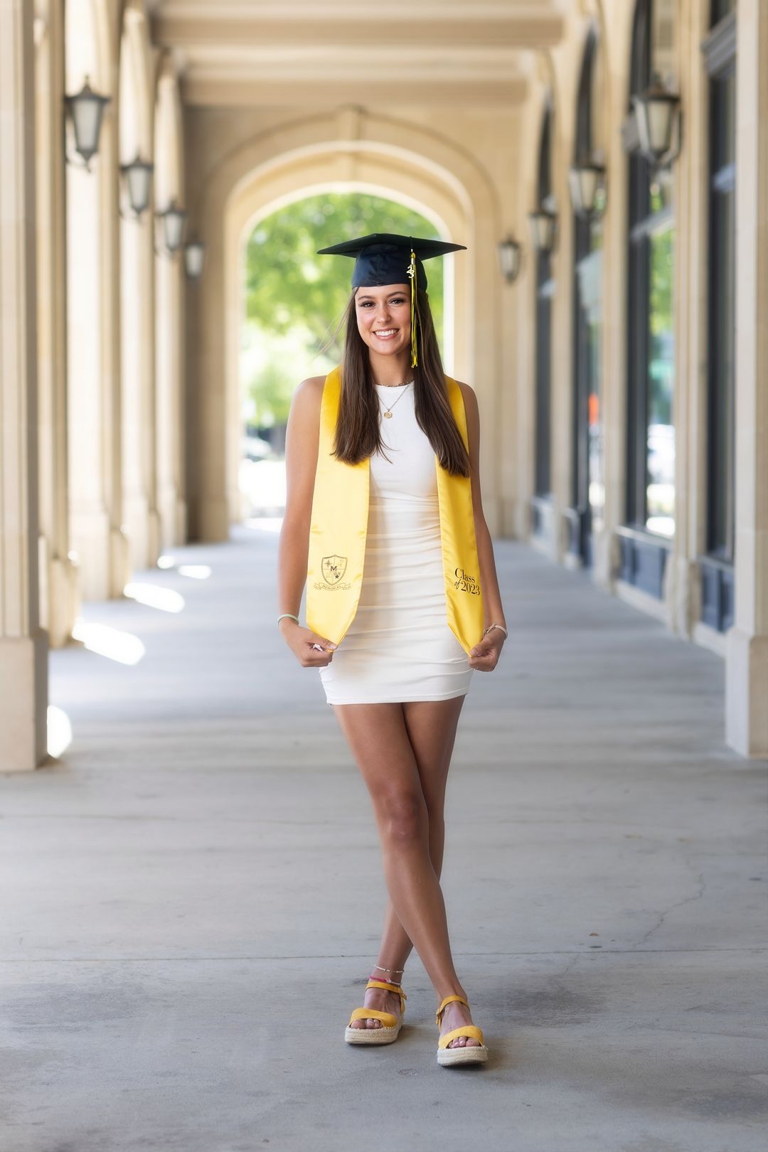 Graduate in cap and gown poses for graduation photos in an elegant covered walkway with arched columns.