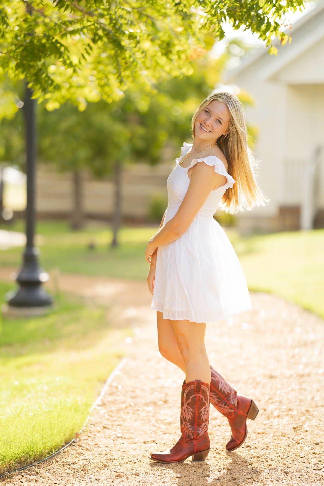 A person in a white summer dress and brown cowboy boots poses playfully on a sunlit path under green trees.