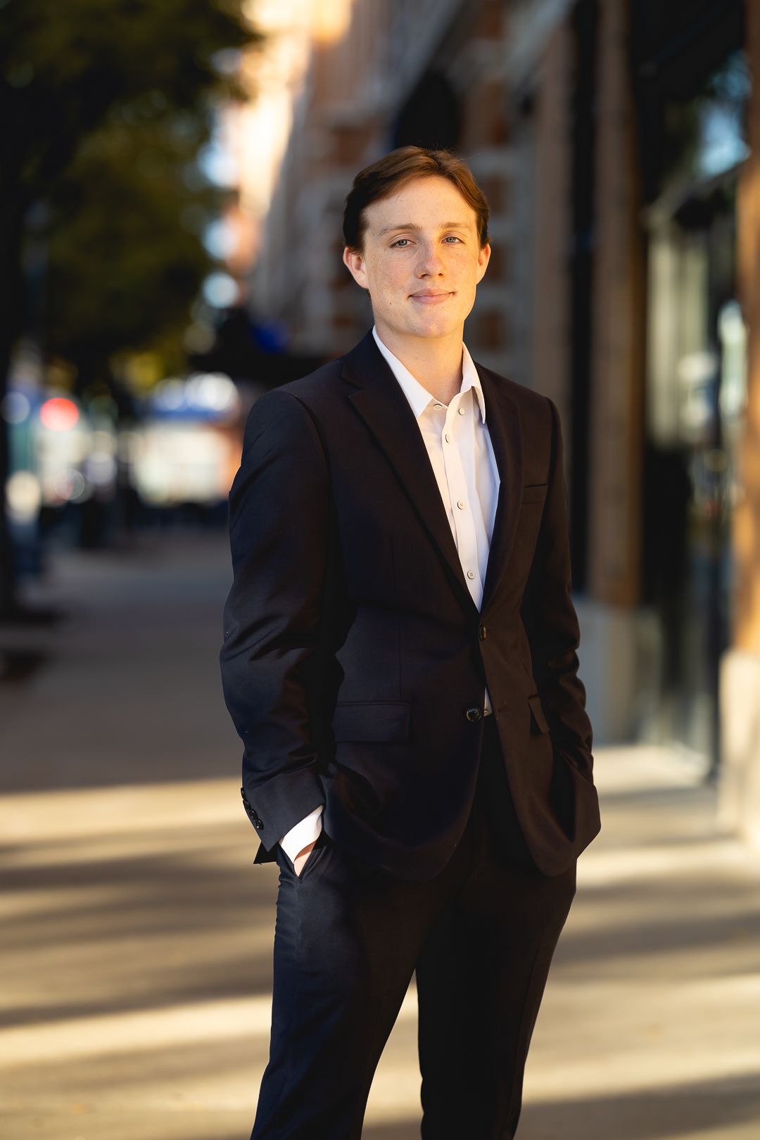 A person in a dark suit jacket stands outdoors with a soft smile against an urban backdrop in warm evening light.