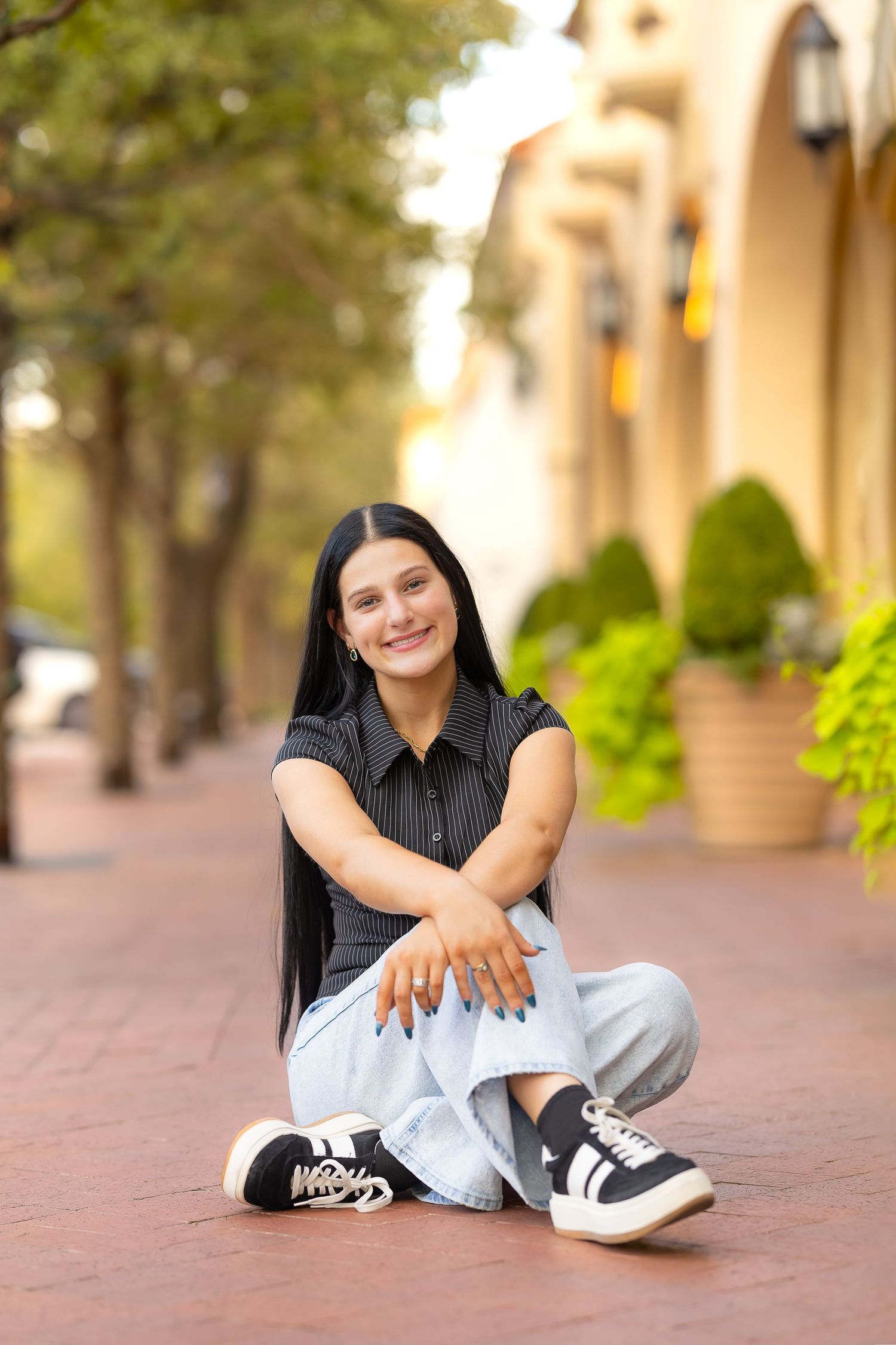 A person in a black top and gray pants sits casually on a brick walkway with trees and buildings in the background.