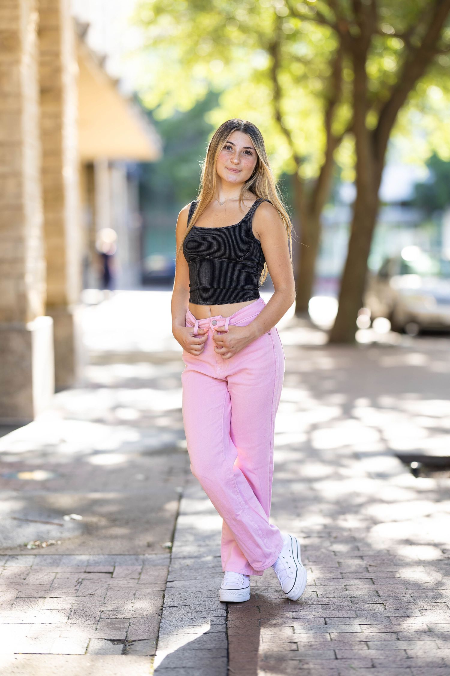 Person wearing black top and pink pants poses in outdoor setting with sunlit trees and brick pavement.