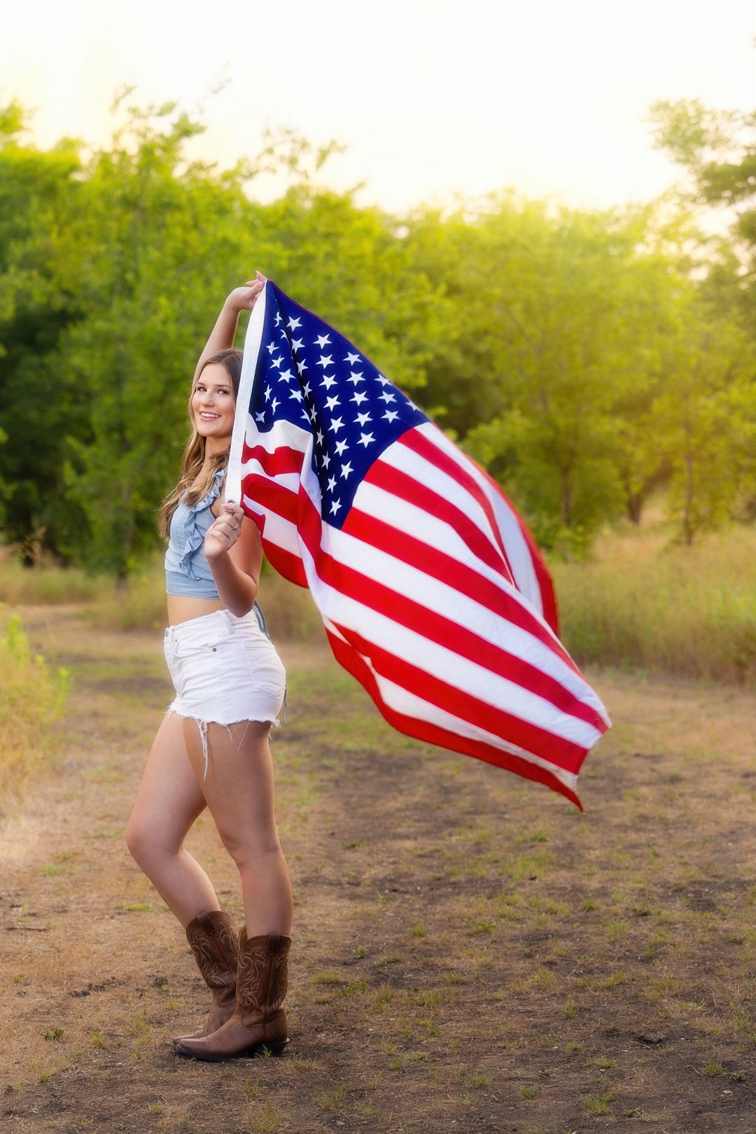 Young woman in denim jacket and white shorts holds American flag outdoors in golden sunlight wearing cowboy boots.