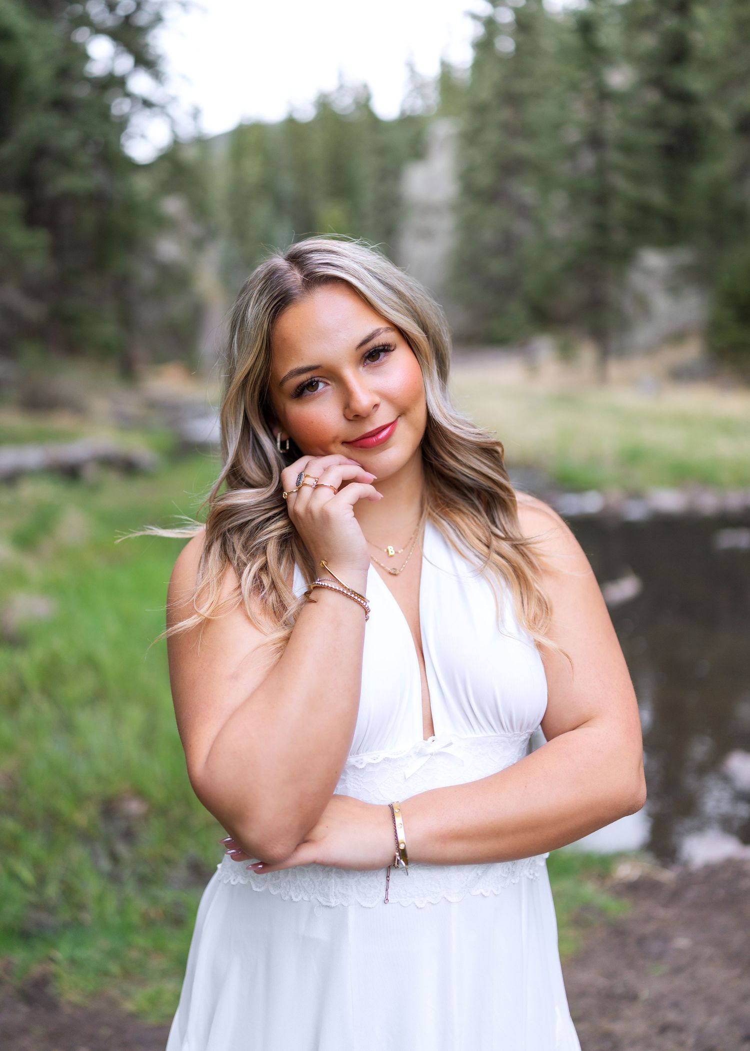 Young woman in white halter dress poses outdoors near a creek surrounded by pine trees.