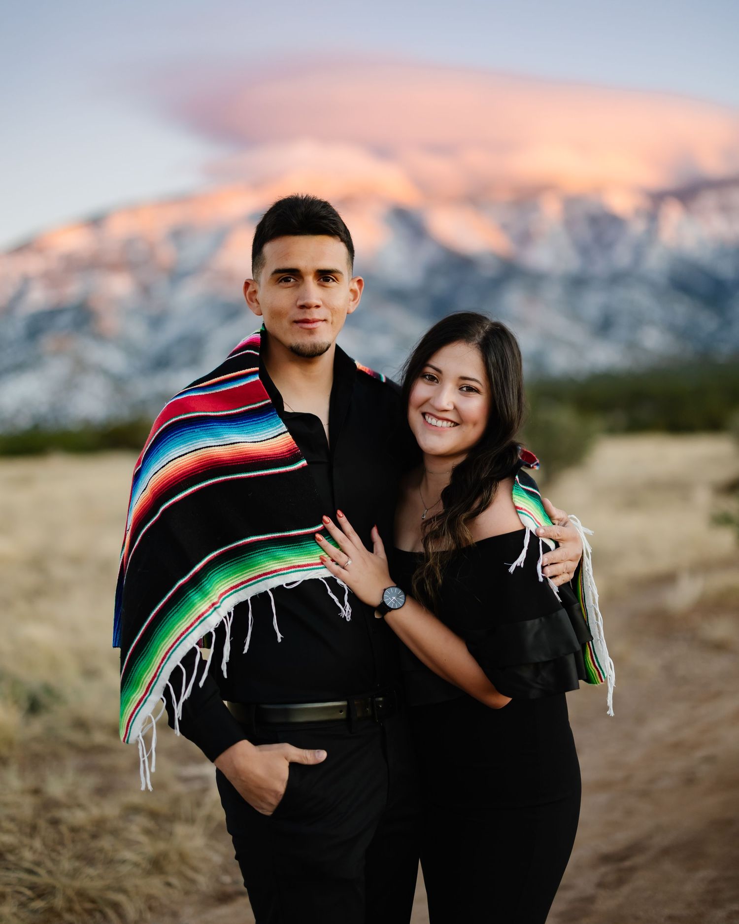 A couple in dark clothing shares a colorful Mexican serape blanket against a snowy mountain backdrop at sunset.