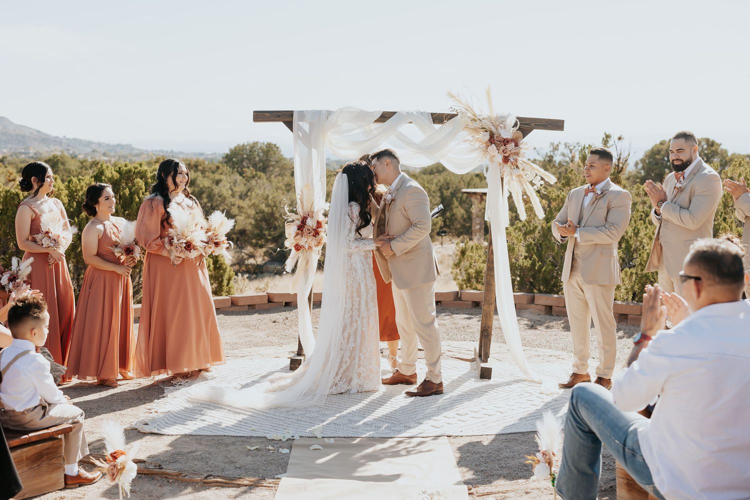 Wedding ceremony attendees gather on a sandy beach during a sunset celebration with desert landscaping.