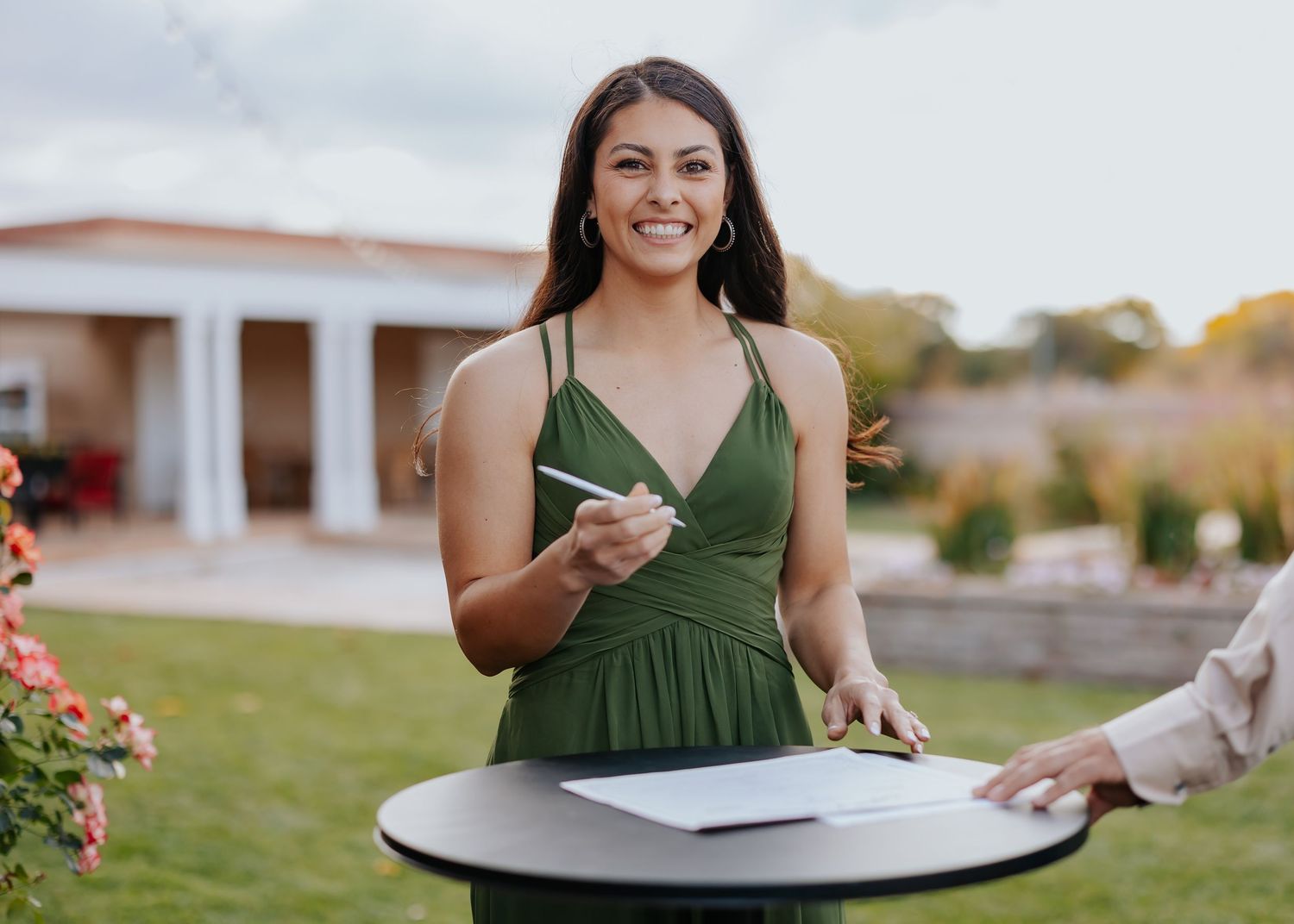 A person in a green dress takes notes while sitting at an outdoor patio table with blooming flowers nearby.