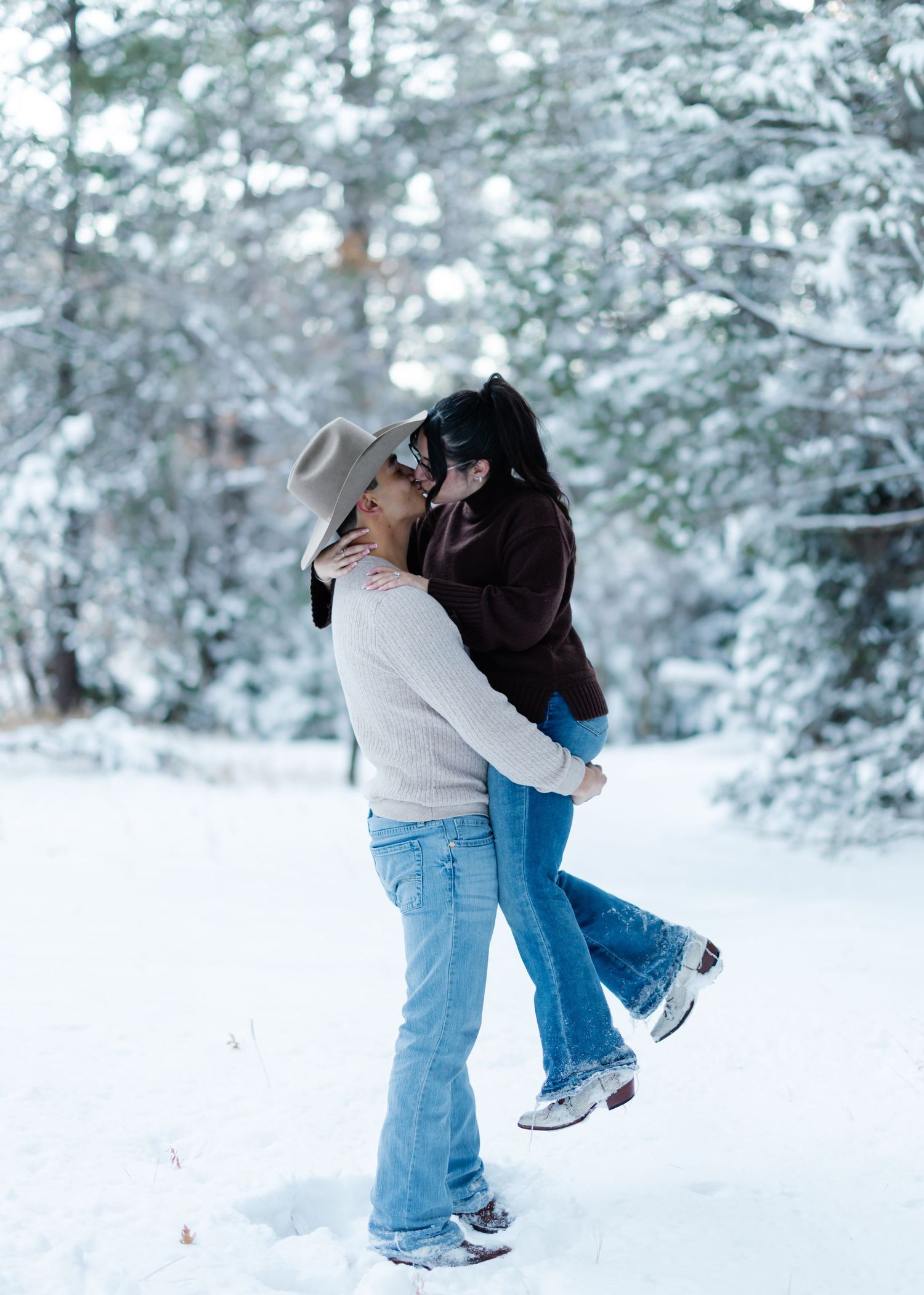 Couple kissing and embracing in a snowy winter forest, wearing jeans and cozy sweaters.