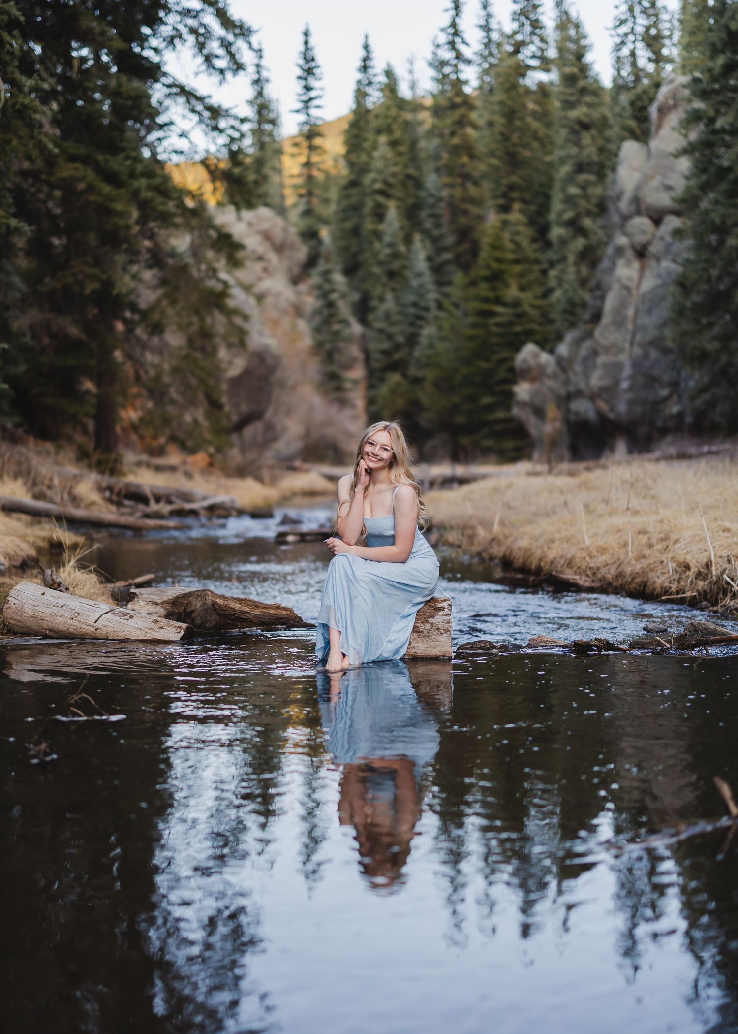 A woman in a light blue gown sits on rocks in a serene mountain stream surrounded by pine trees and rocky cliffs.