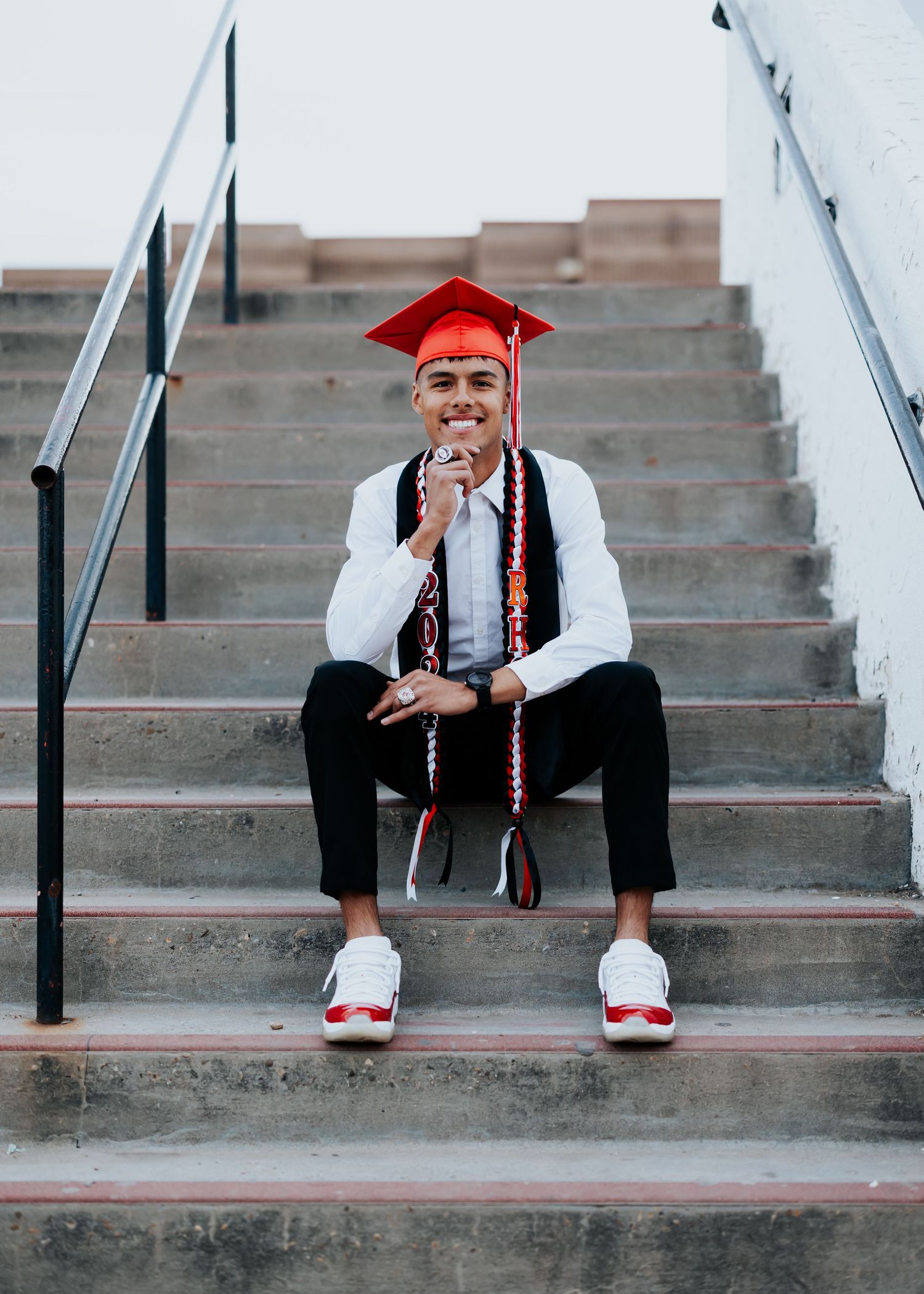 Graduate in red cap and gown sits on concrete steps for casual portrait series.
