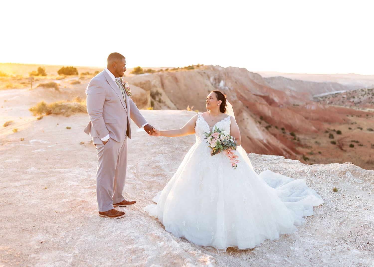 Bride and groom hold hands at sunset on a rocky desert landscape, bride in a ballgown with bouquet.