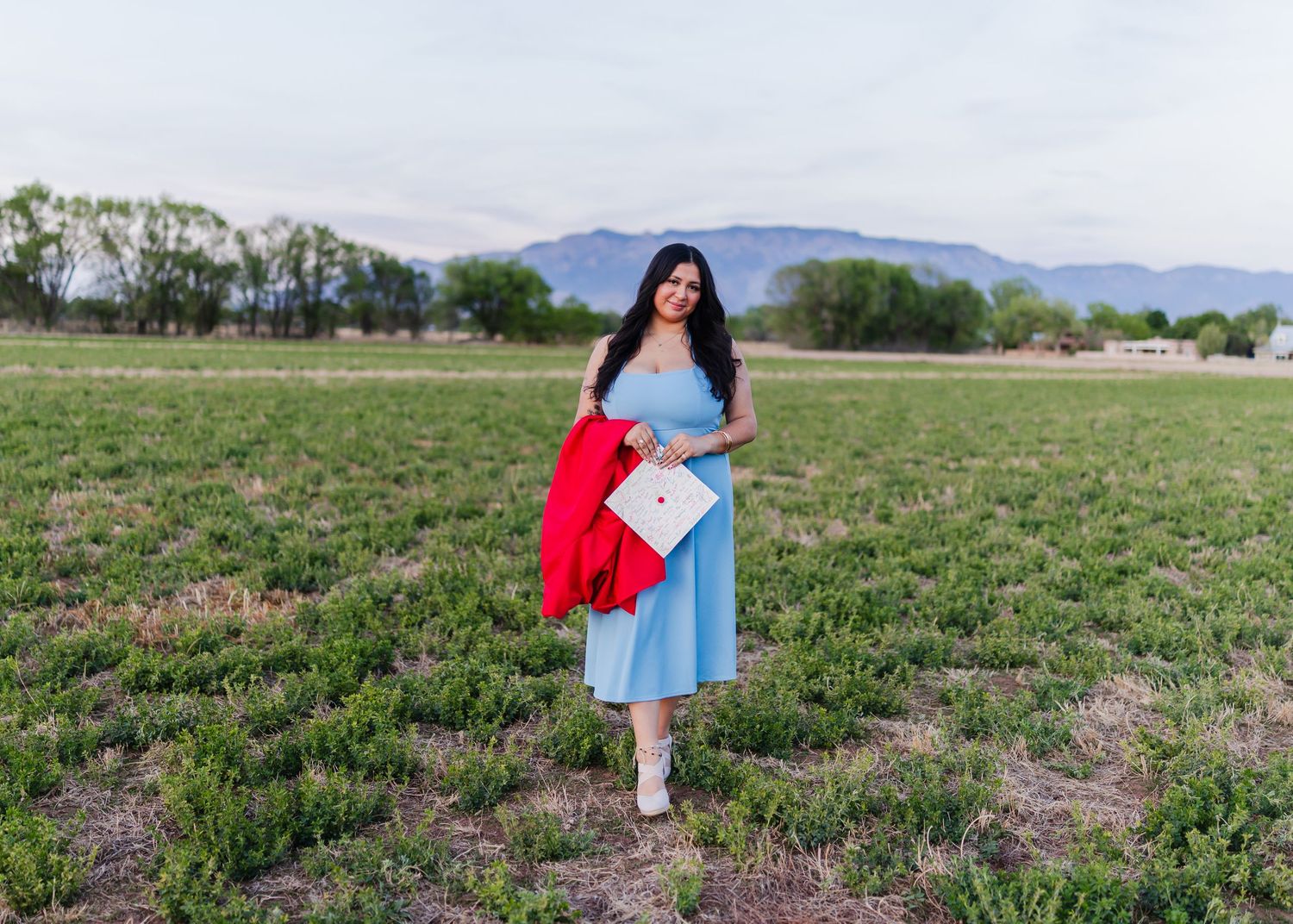 Woman in blue dress holding red graduation gown and cap standing in green field with mountains backdrop.
