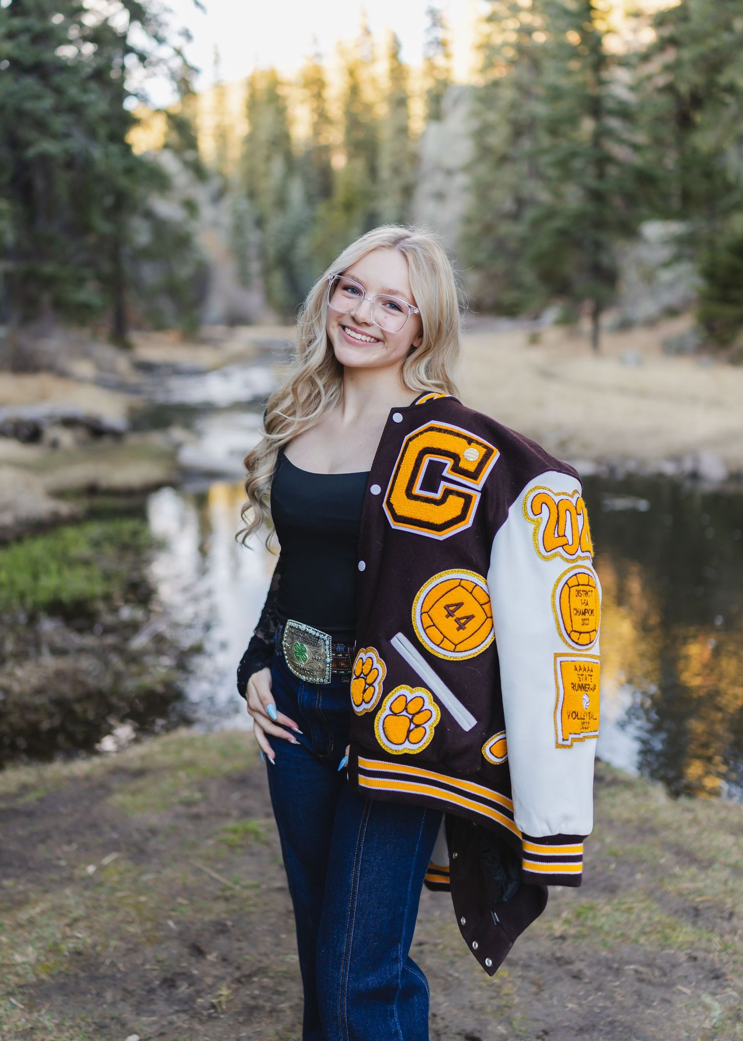 Smiling blonde teen wearing a varsity letterman jacket with patches poses outdoors near a creek surrounded by pine trees.