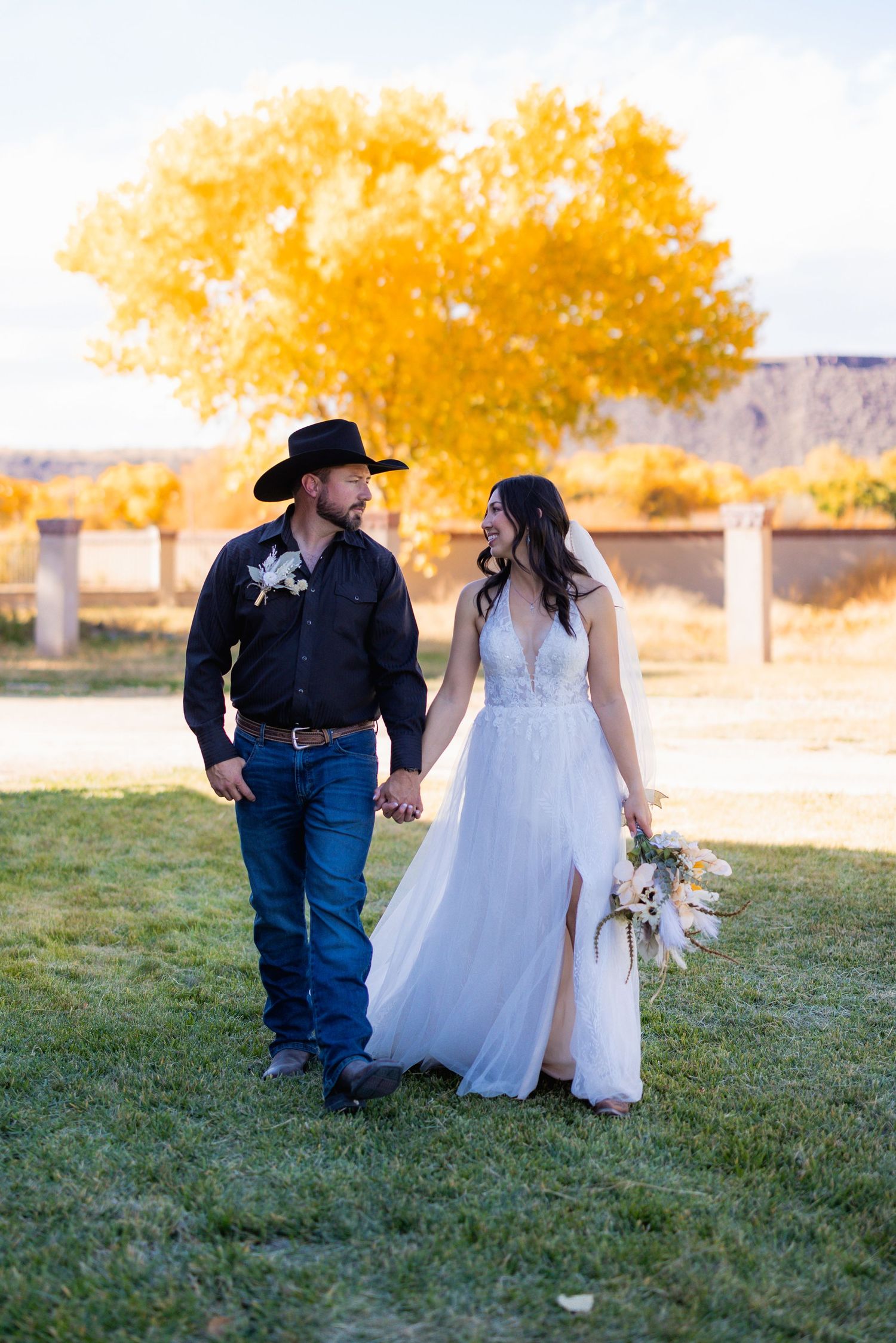 A couple in western wedding attire holds hands while walking on grass with golden autumn trees in the background.