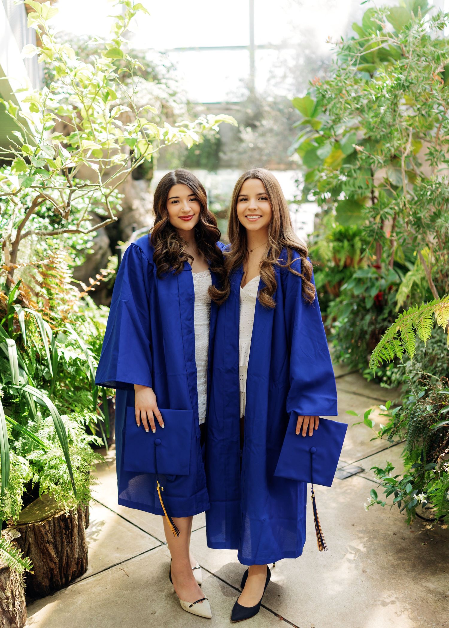 Two young women in blue graduation gowns and caps smiling in a lush greenhouse setting.