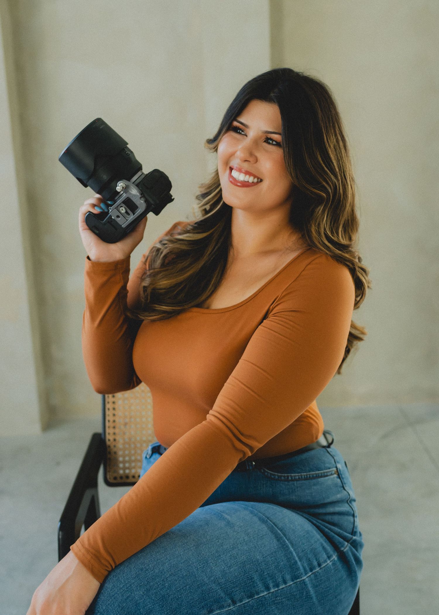 Woman in rust-colored long sleeve top and jeans poses casually with coffee cup against neutral wall.