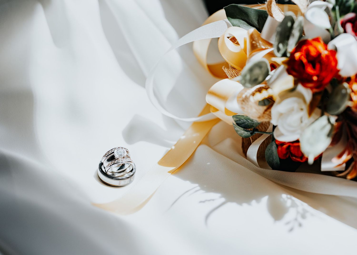 A platinum wedding band and engagement ring rest on a white silk fabric alongside delicate pink and white floral arrangements.