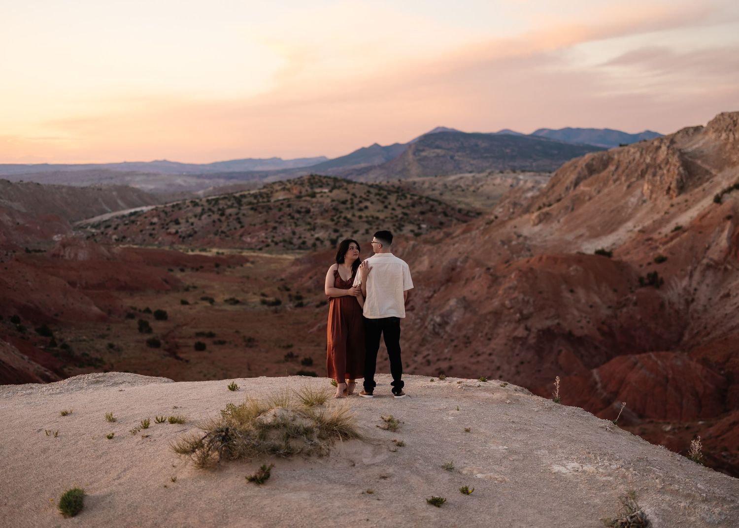 Two silhouettes stand together on a rocky overlook during sunset in a desert landscape.