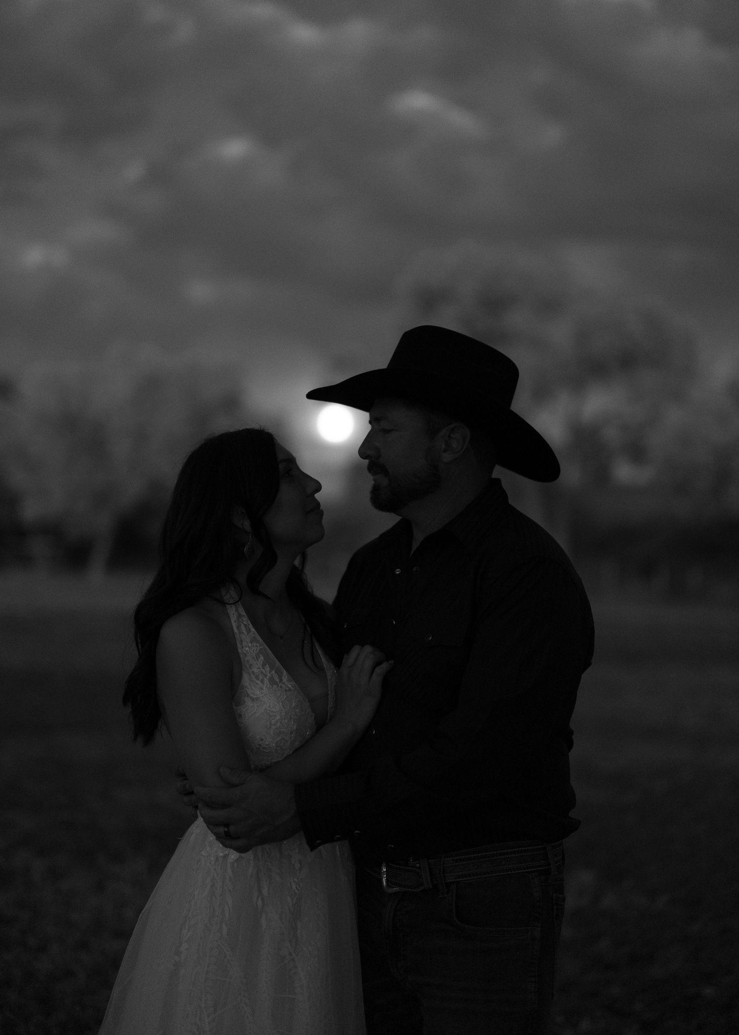 A romantic silhouette of a couple embracing outdoors with the moon glowing behind them in black and white.