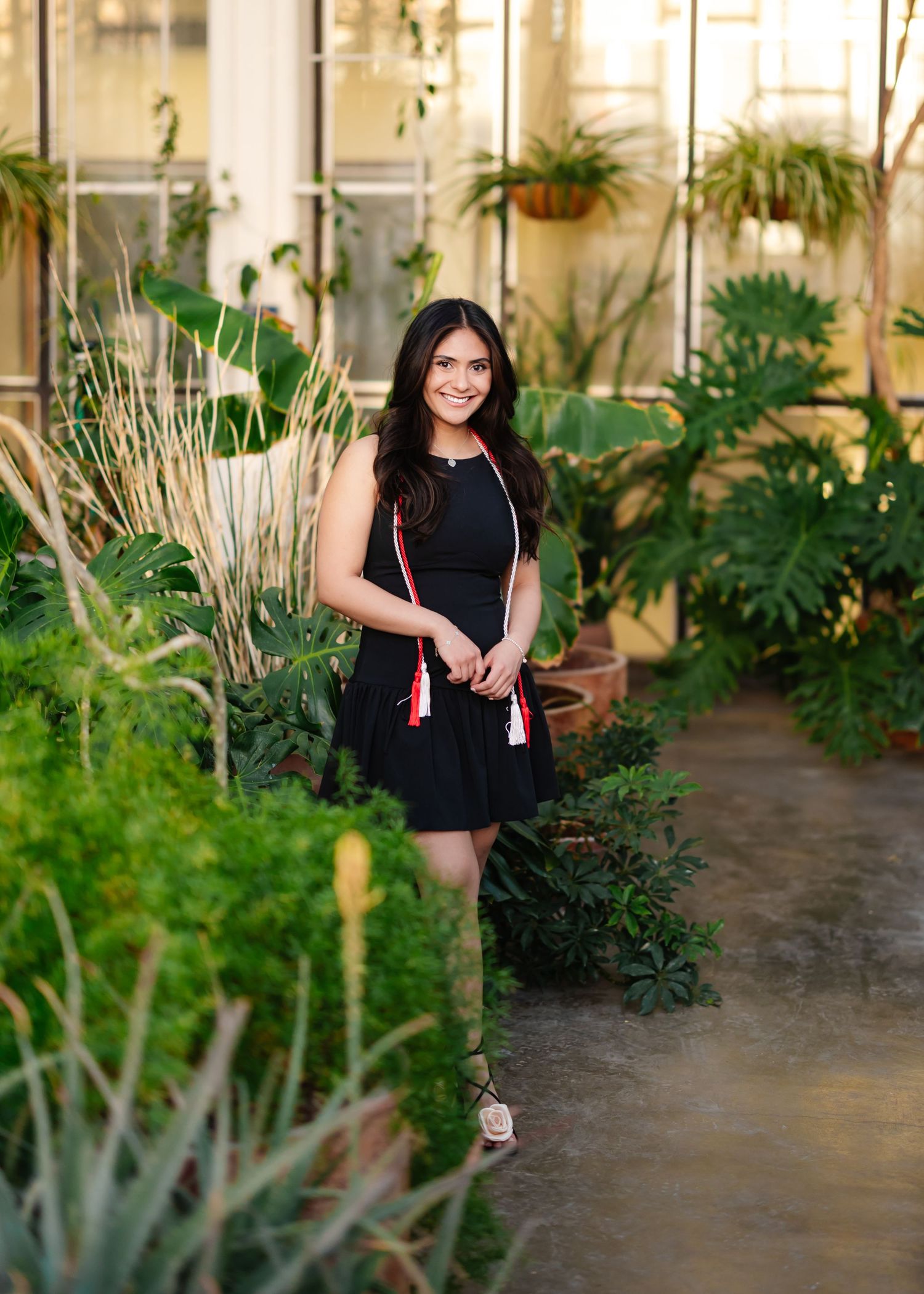 A woman in a black dress stands in a lush indoor garden setting with tropical plants.