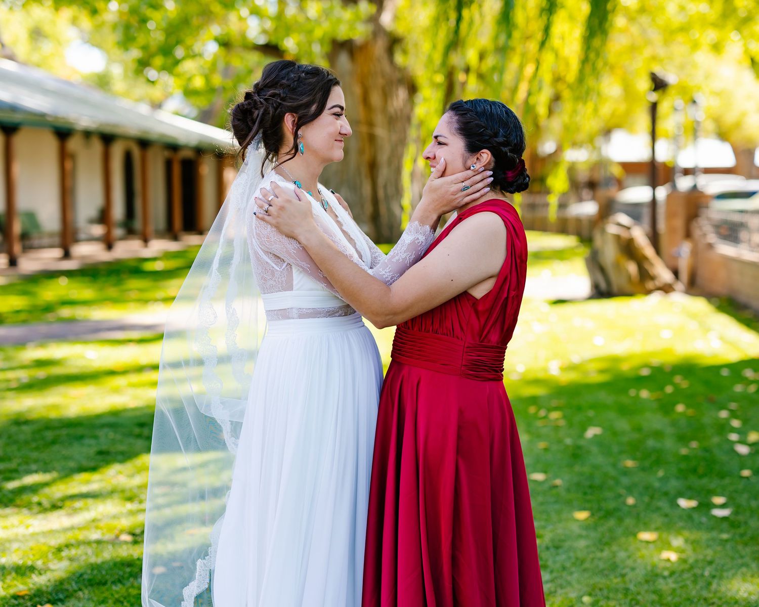 Two people wearing red and white dresses embrace emotionally during an outdoor wedding ceremony on a sunny day.