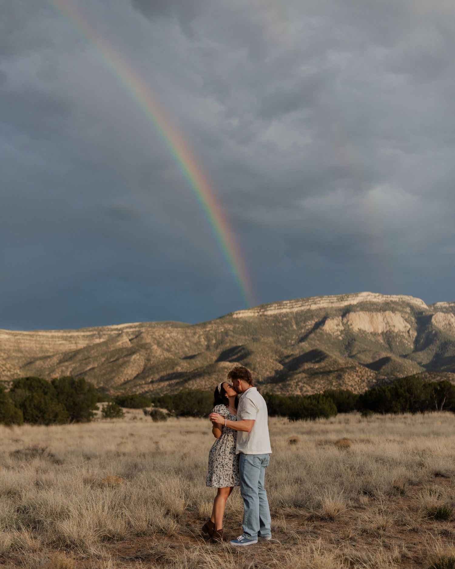 Couple embracing in a field with a vibrant rainbow over dramatic mountains and stormy skies in the background.