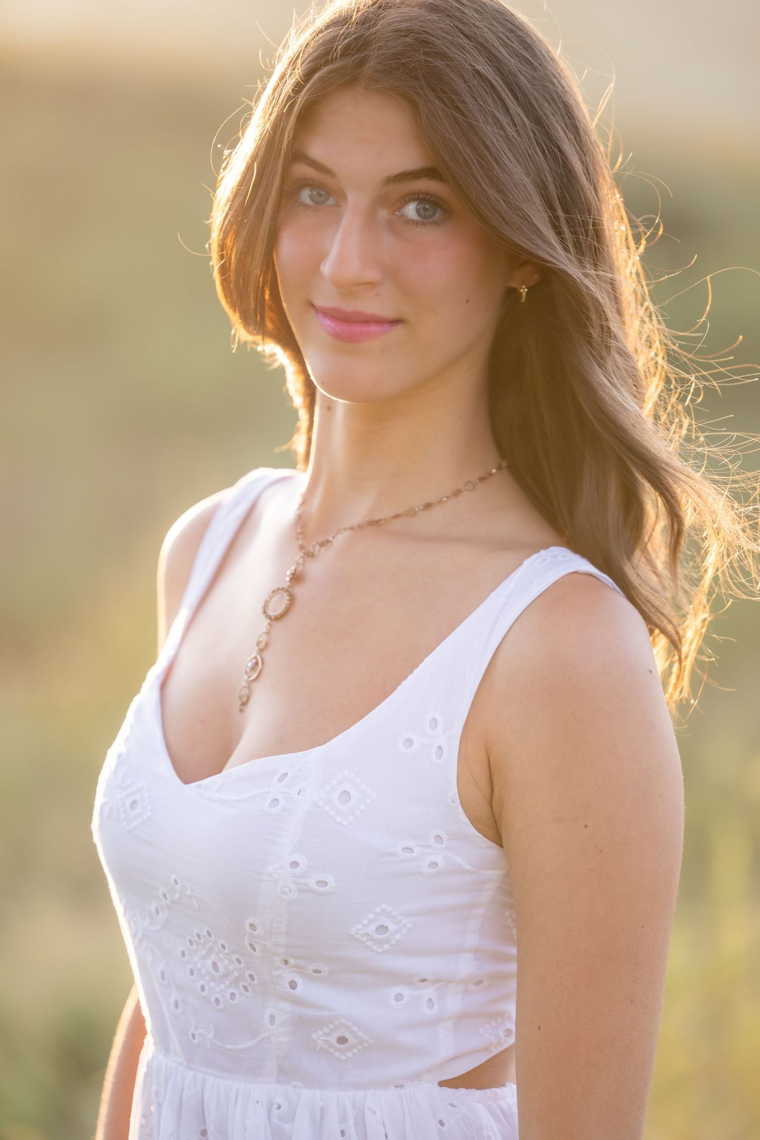 A portrait in golden sunlight shows someone wearing a white summer dress with delicate embroidery and a dainty necklace.
