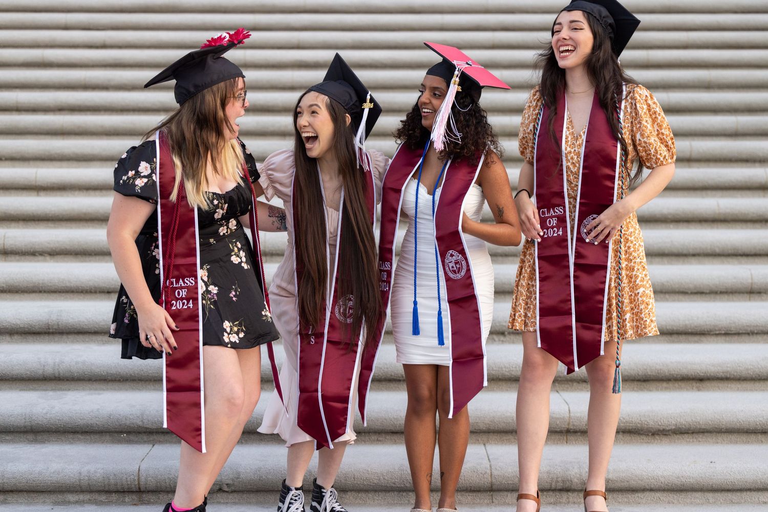 A group of graduates in burgundy gowns and black caps pose together on stone steps during graduation.