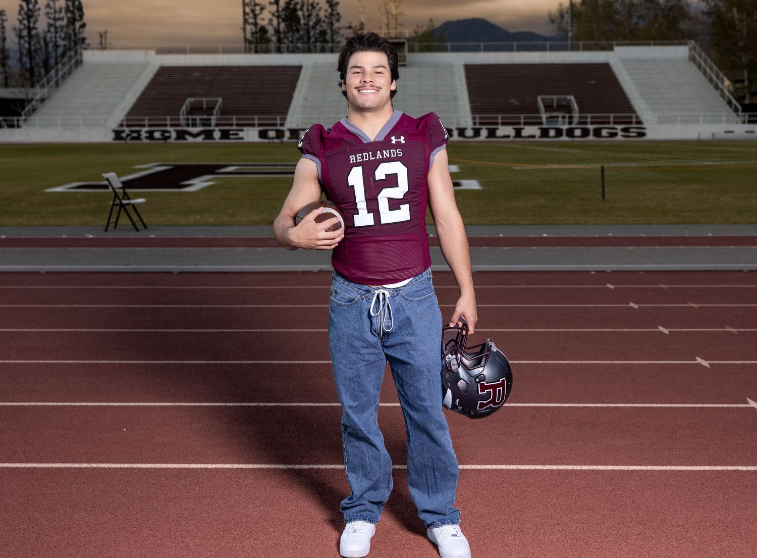 Football player wearing jersey number 12 stands on track field holding helmet and ball at sunset with stadium in background.
