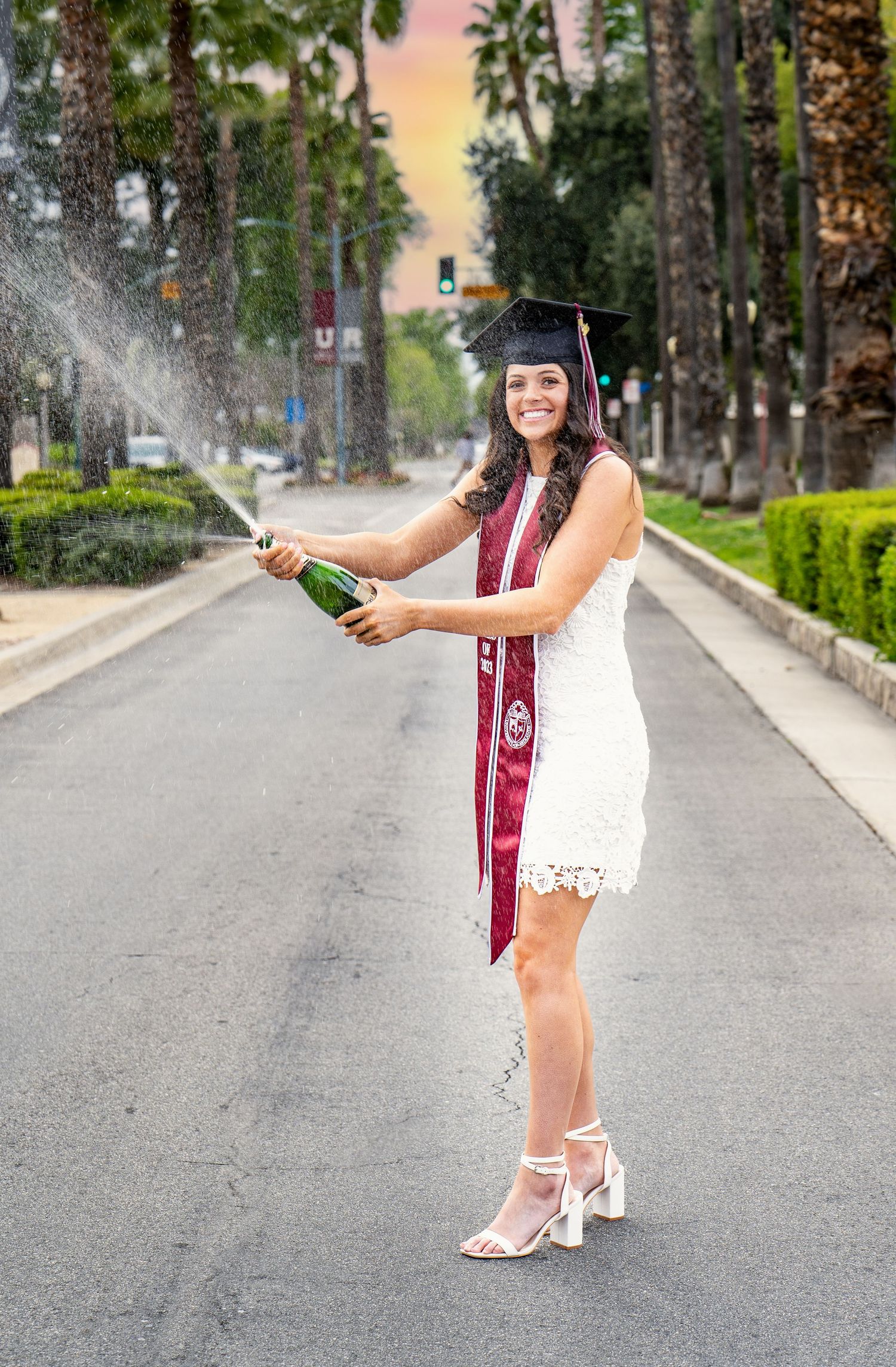 Graduate in white dress and burgundy stole poses on tree-lined street during sunset celebration photoshoot.